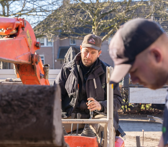 Een arbeider met een bruine pet bedient een oranje graafmachine op een zonnige bouwplaats, met een collega in de buurt.