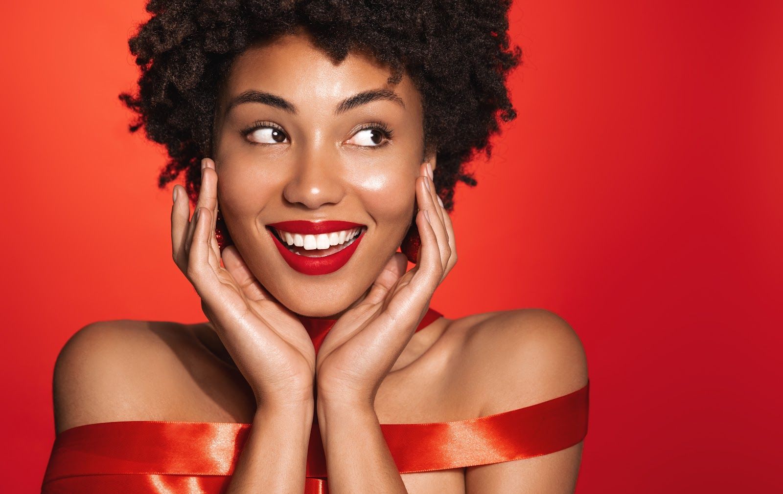 Smiling woman in a red off-shoulder dress against a bright red background, hands framing her face