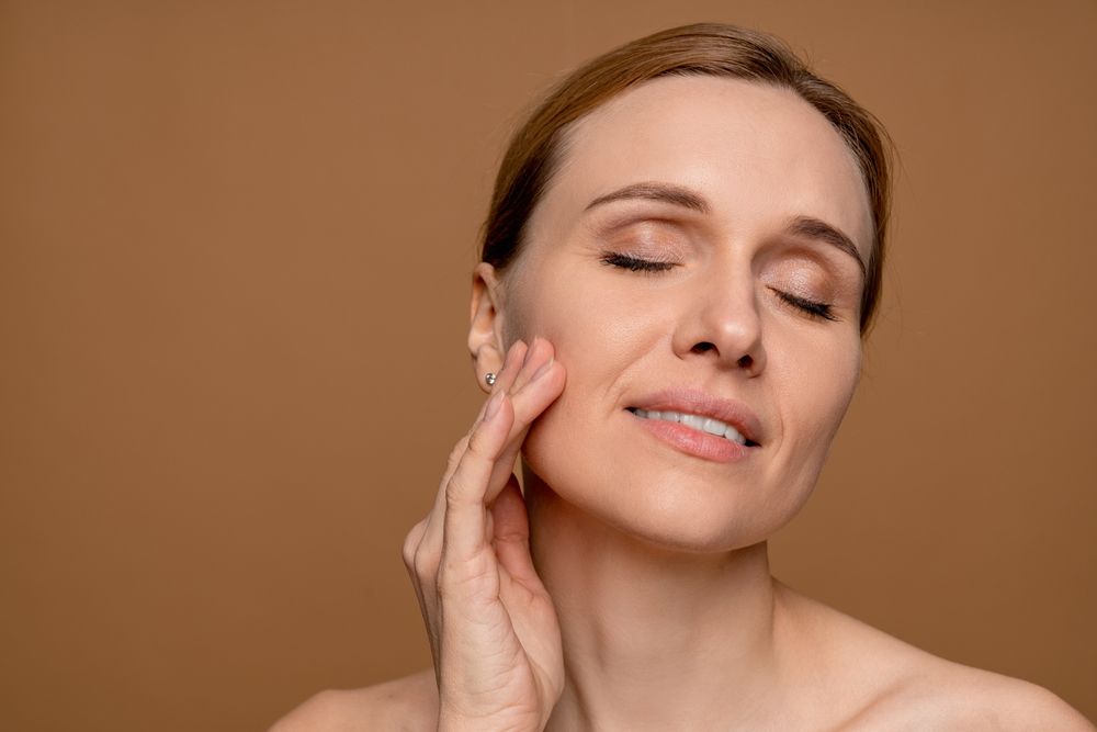 A person with eyes closed touches their cheek with one hand against a plain brown background.