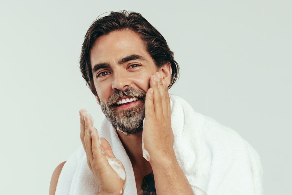 A bearded person with a towel over their shoulders smiling while applying cleansing foam to their face.