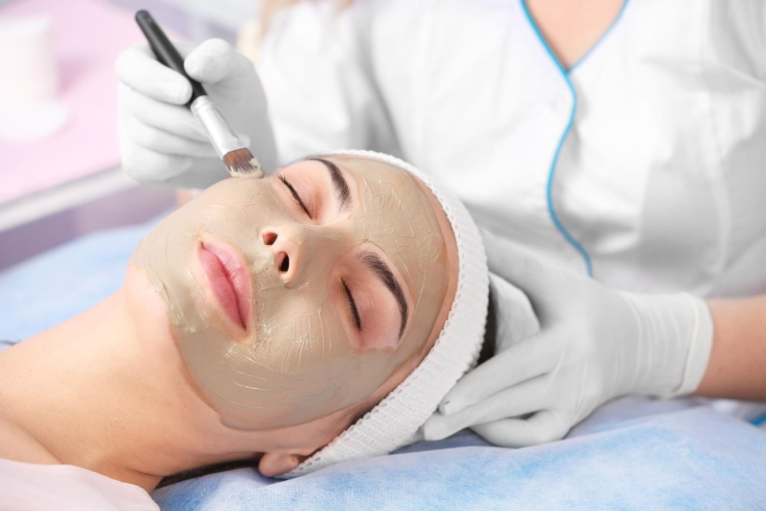 A person wearing a white headband receives a facial treatment with a beige mask applied by a gloved aesthetician.