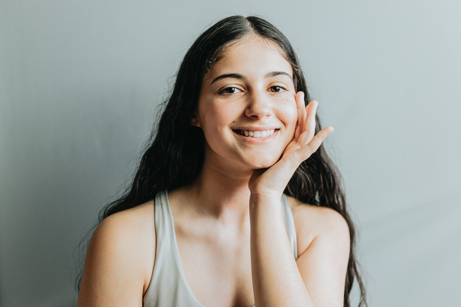 Smiling person with long dark hair rests their face on their hand against a gray background