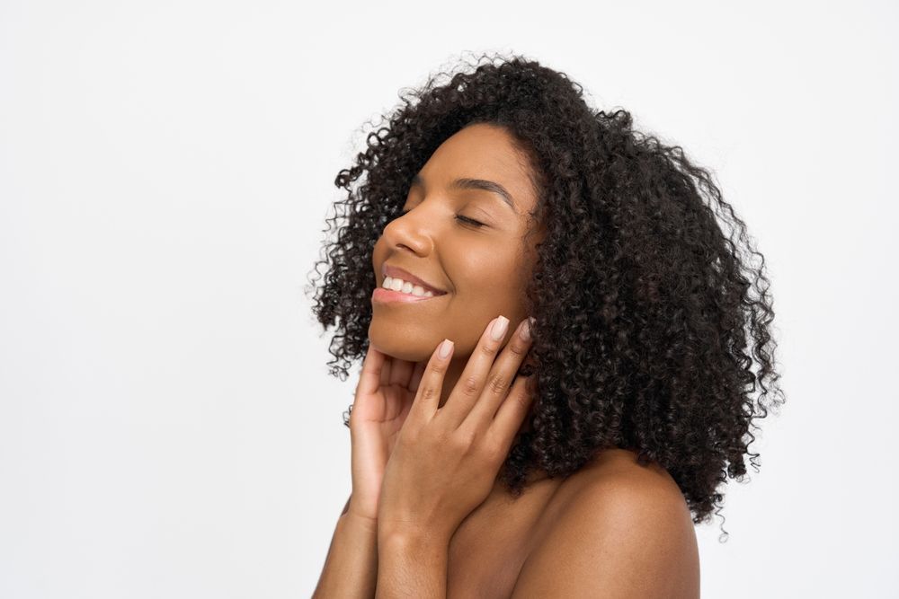 A person with curly hair, eyes closed, and a gentle smile, holding their hands against their face against a white backdrop.