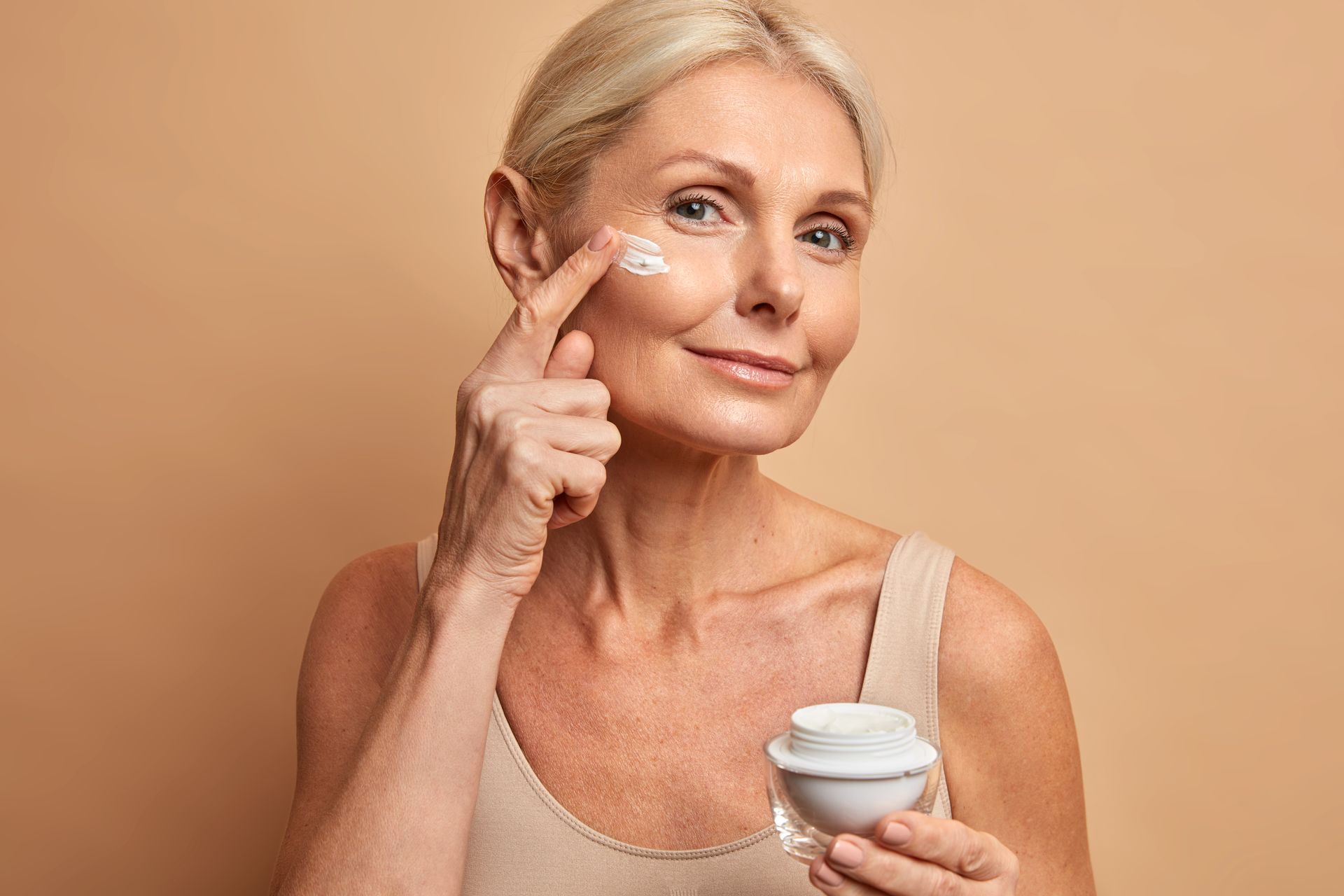 A woman is applying cream to her face while holding a jar of cream.