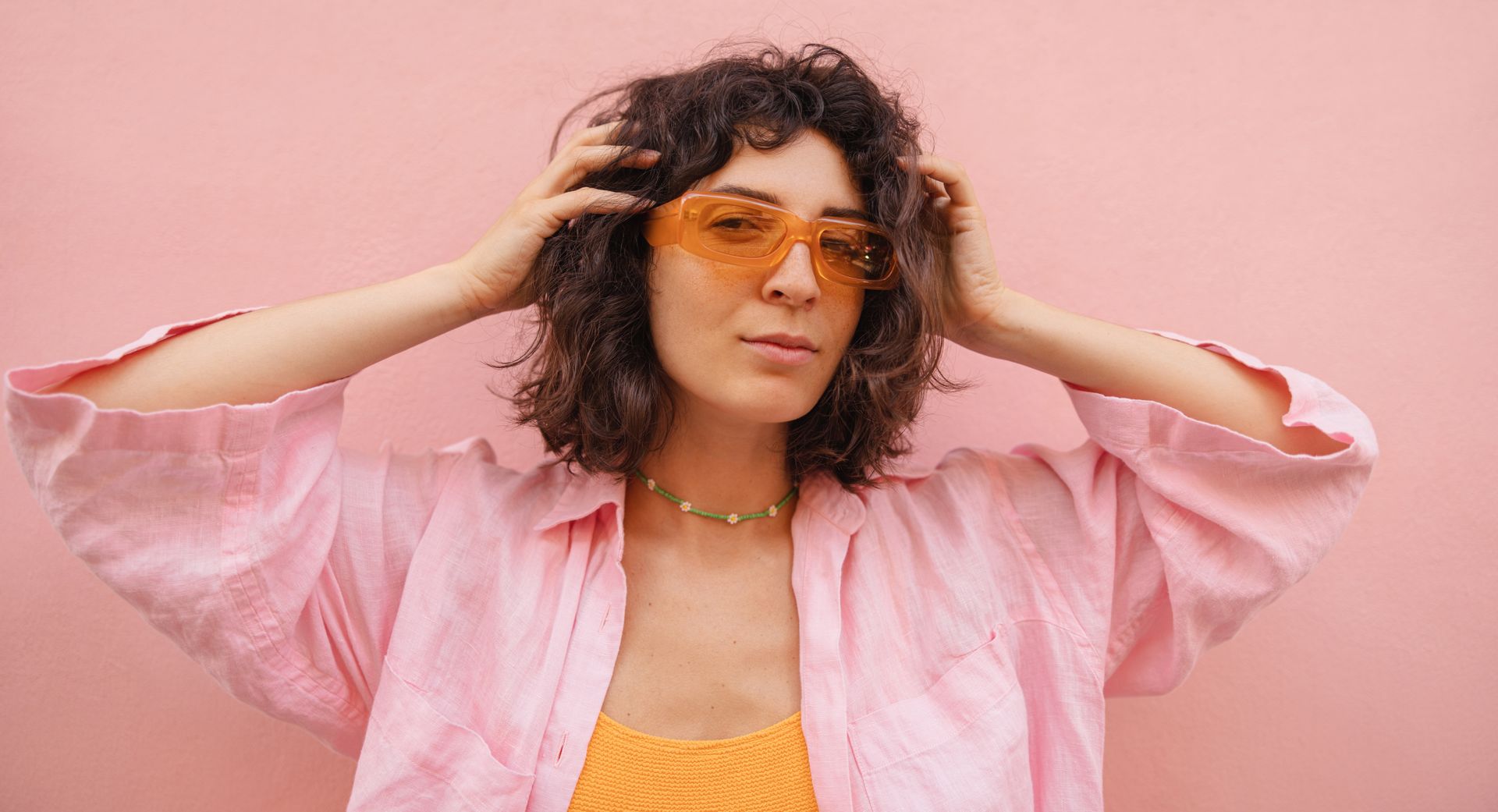 Person in orange-tinted glasses and pink shirt posing against a pink background.