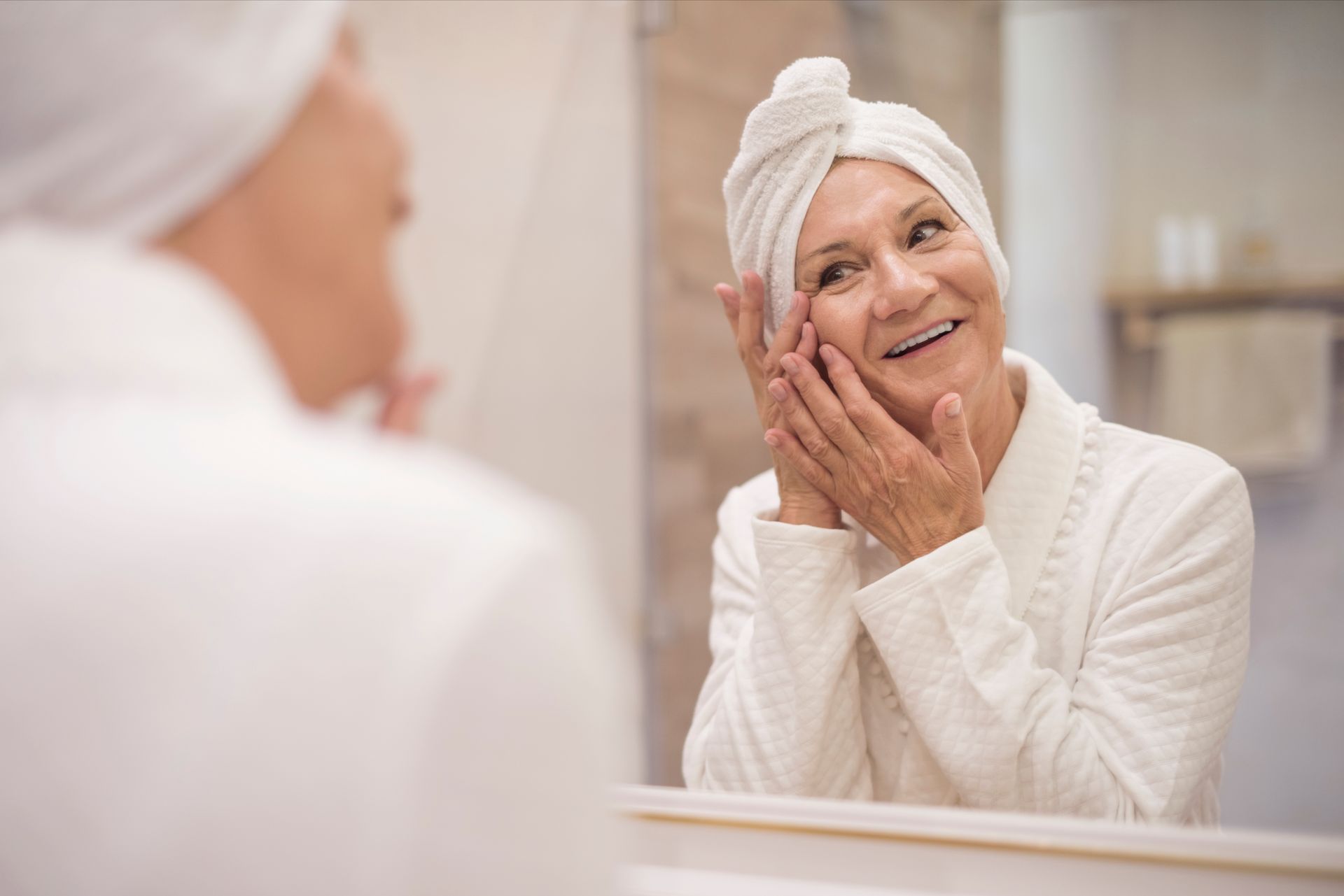 An elderly woman with a towel wrapped around her head is looking at her face in the mirror.