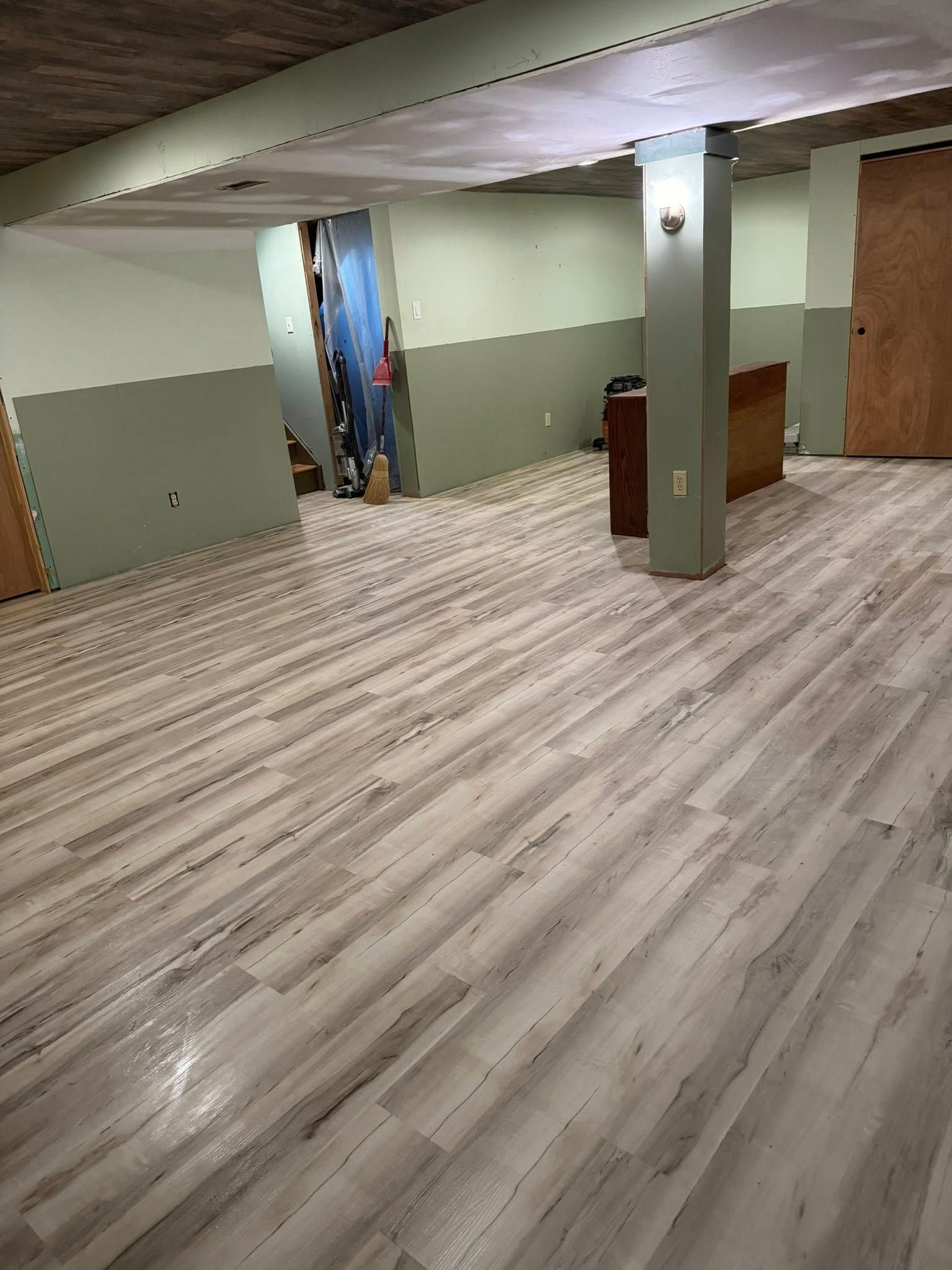 Finished basement with light wood-look flooring and light green walls; concrete ceiling, wooden doors, and supporting column.