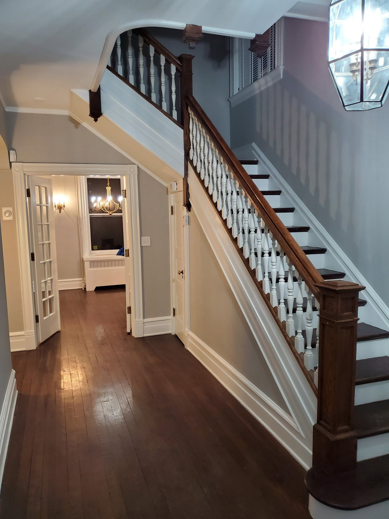 A home's entryway with wooden floors, stairs, and a chandelier. The walls are gray and a doorway is on the left.