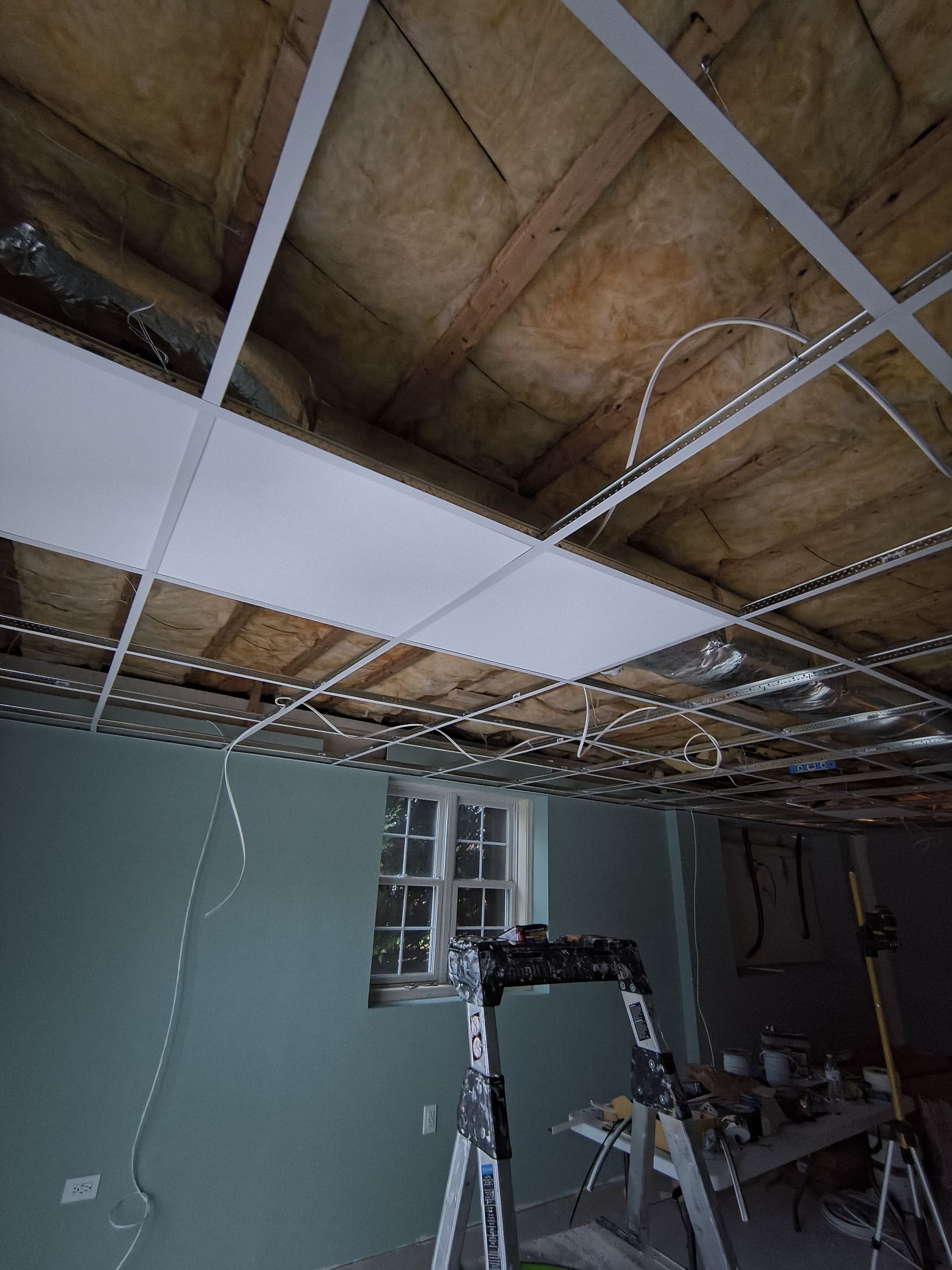 Ceiling tile installation with ladder, exposed wood, wiring, and a window in a room with blue-green walls.