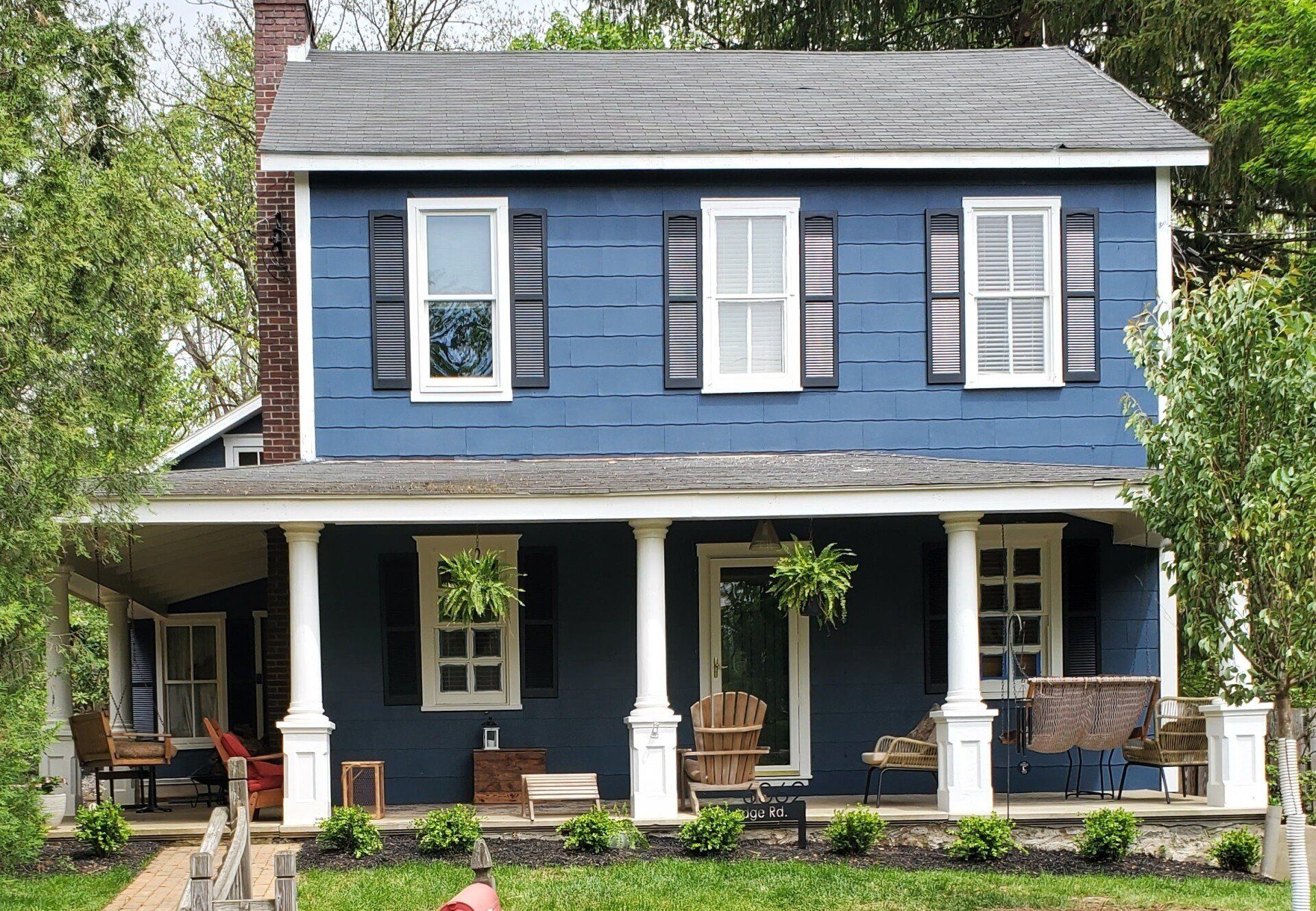 Two-story blue house with white trim, porch, black shutters, and greenery.