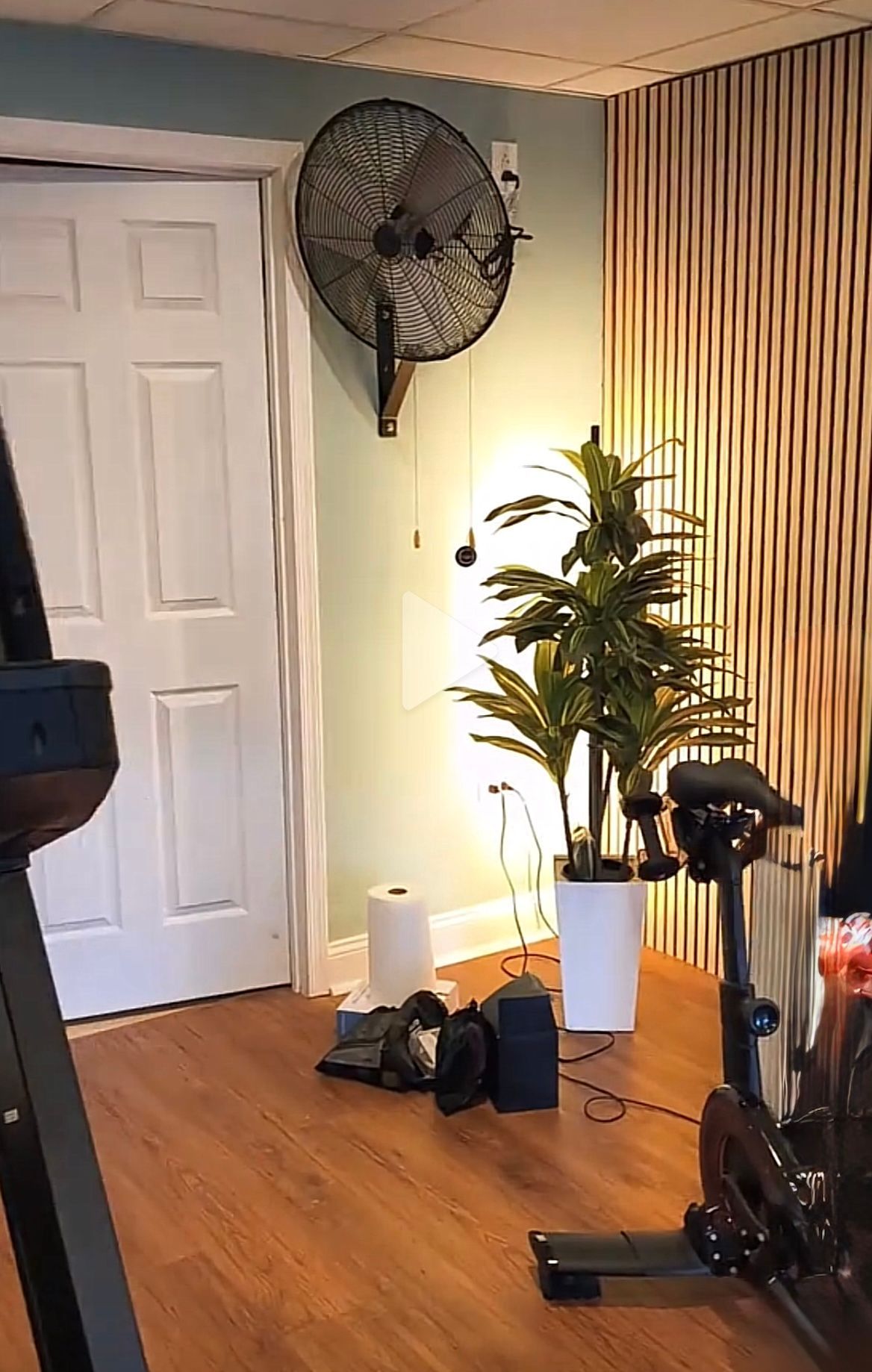 A home gym with a fan, plant, spin bike, and exercise equipment. Light brown walls and wooden flooring.