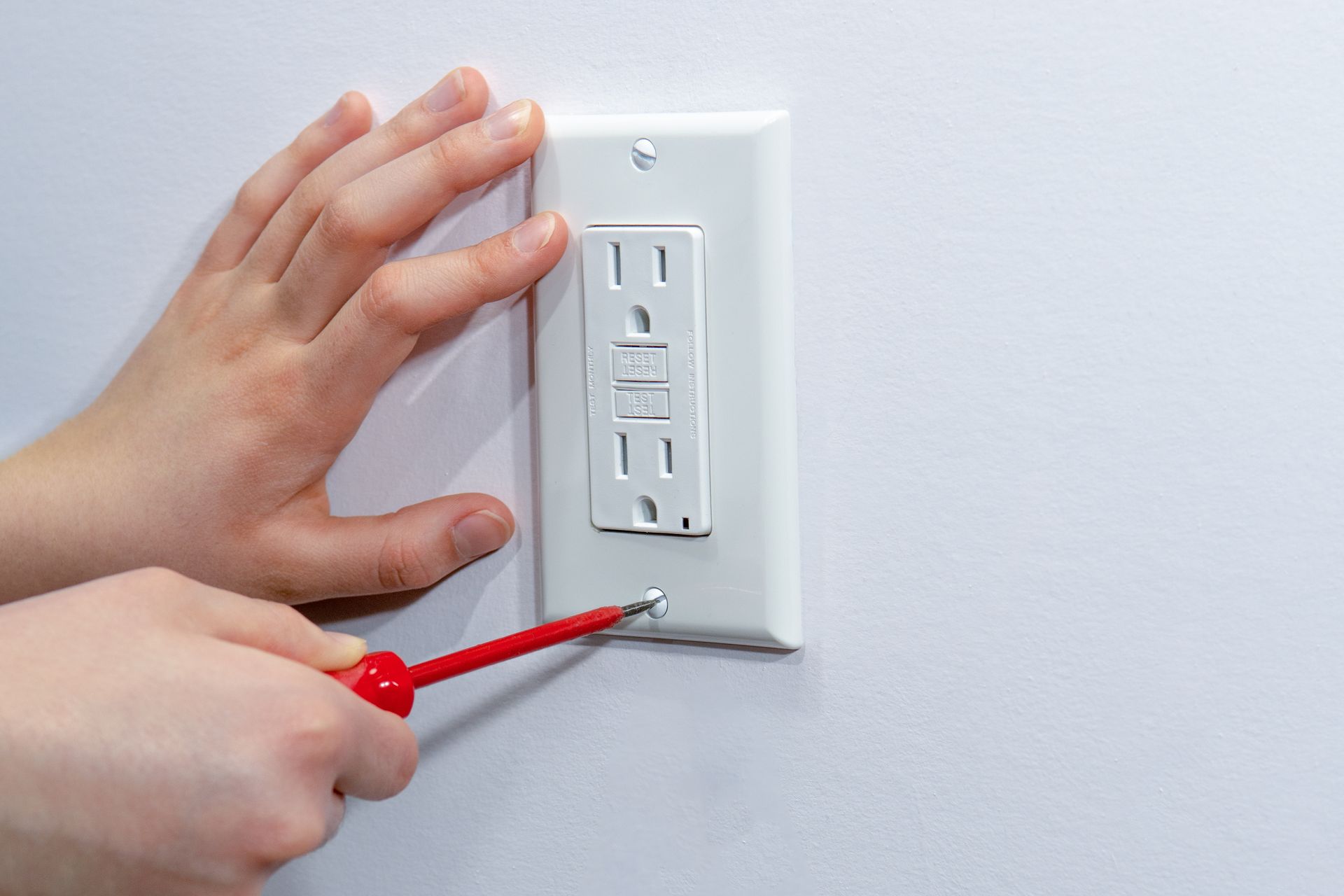 Person using a red screwdriver to tighten a screw on a white electrical outlet cover on a wall.