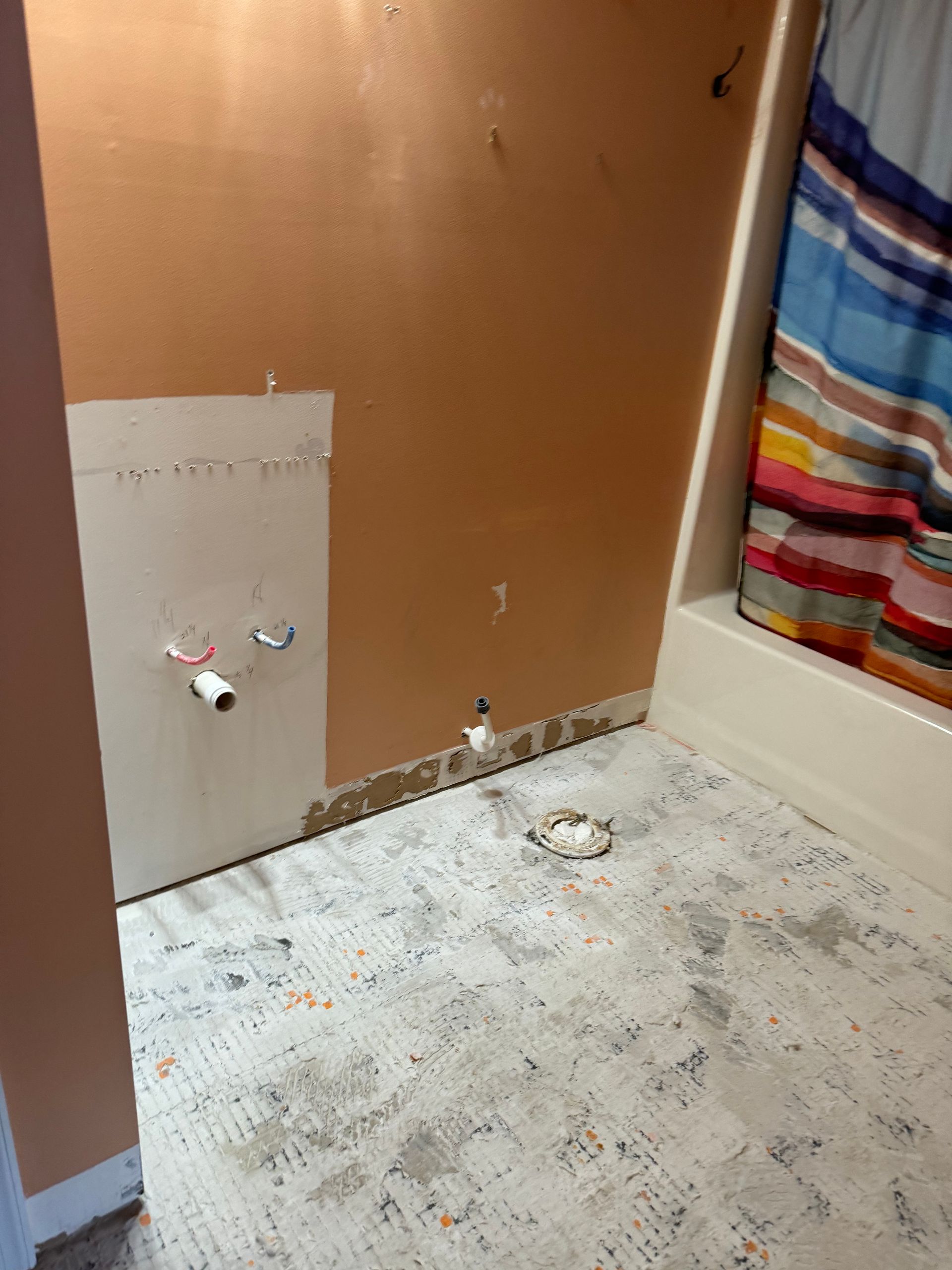 Bathroom renovation. Orange walls, exposed pipes, and damaged flooring; a shower curtain on the right.