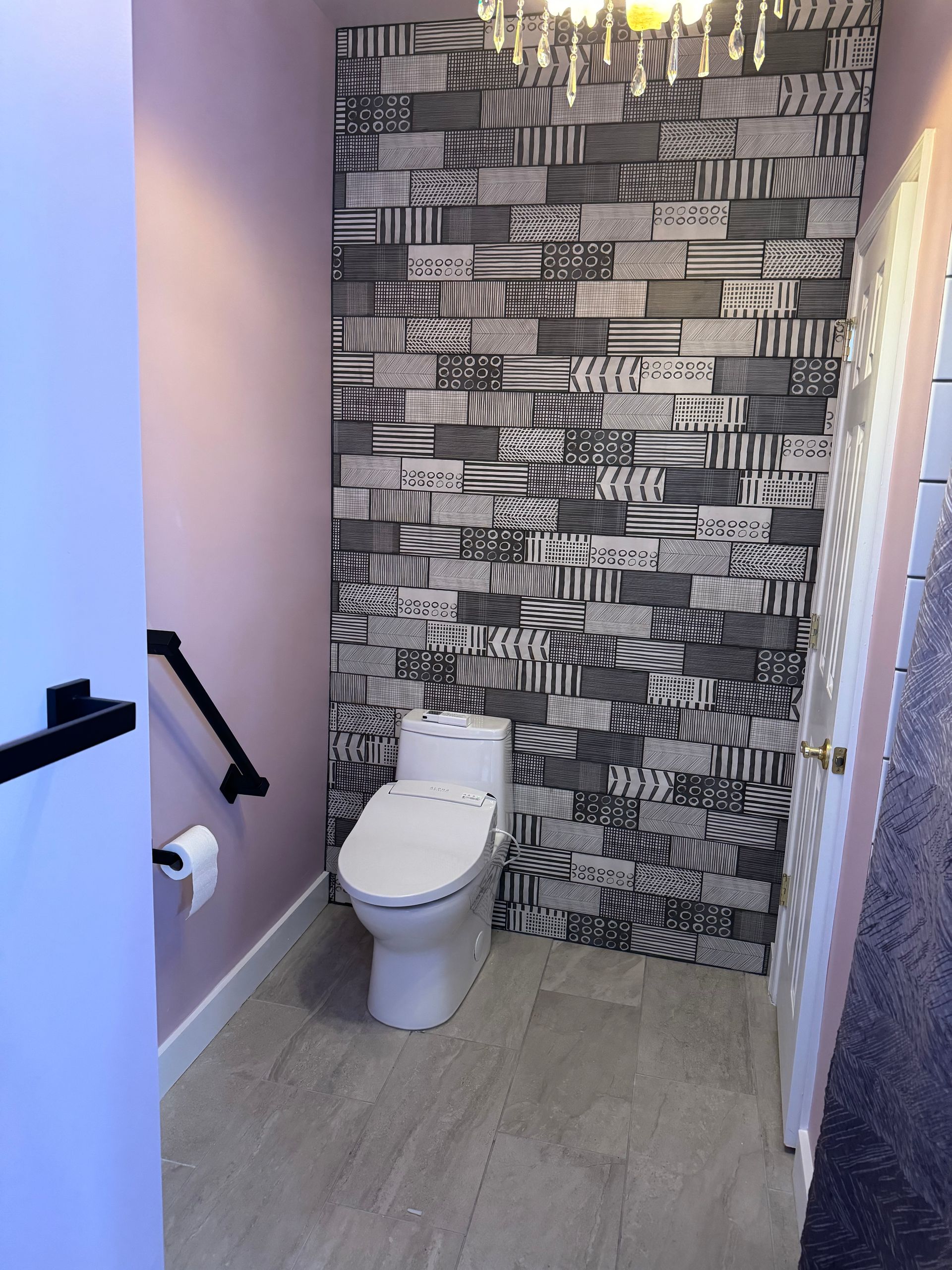 Bathroom with patterned accent wall, white toilet, and gray wood-look flooring. Pink walls and black grab bar.