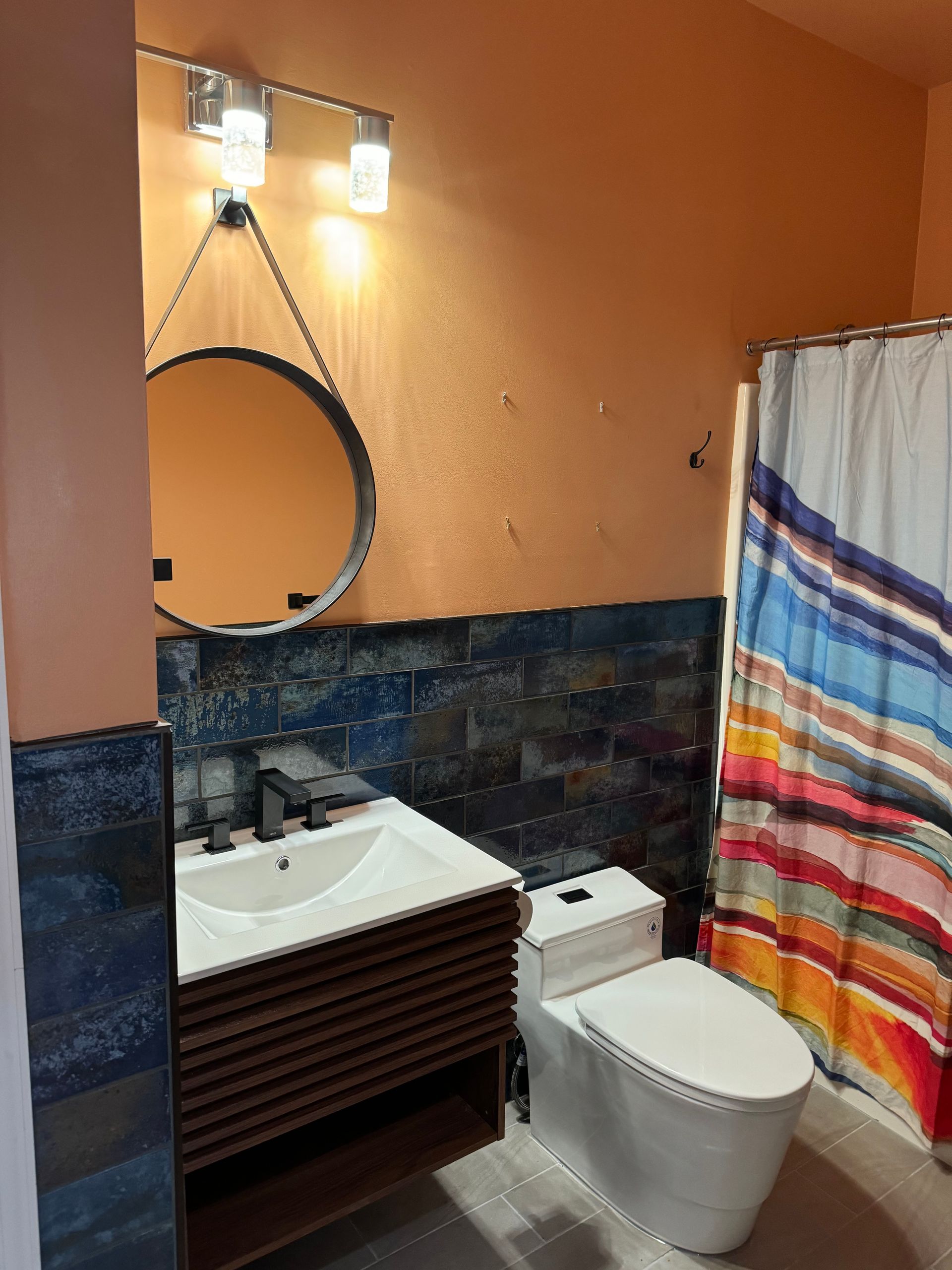 Bathroom with orange walls, blue tile, white toilet, vanity, and a colorful striped shower curtain.
