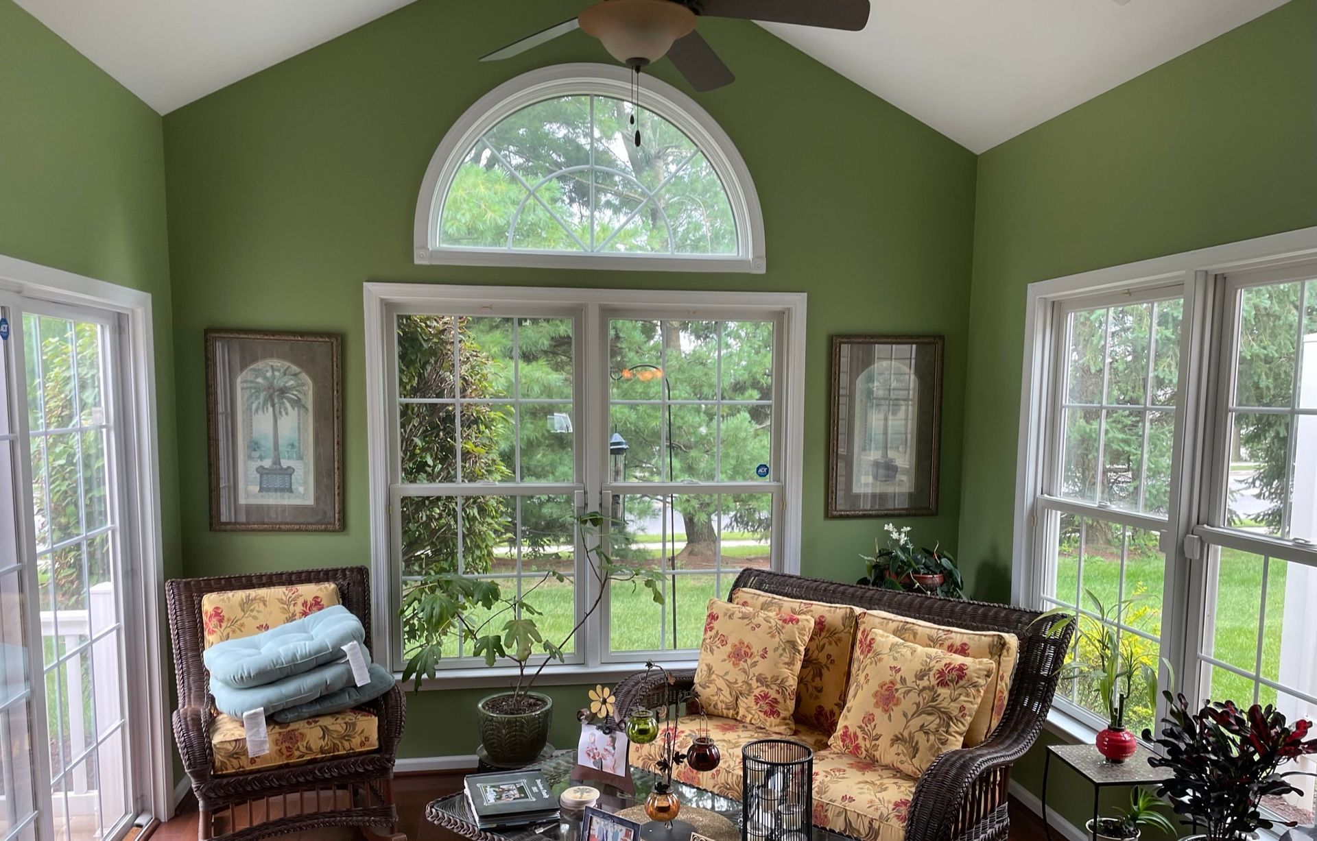 Sunroom with green walls, wicker furniture, and large windows overlooking a green yard.