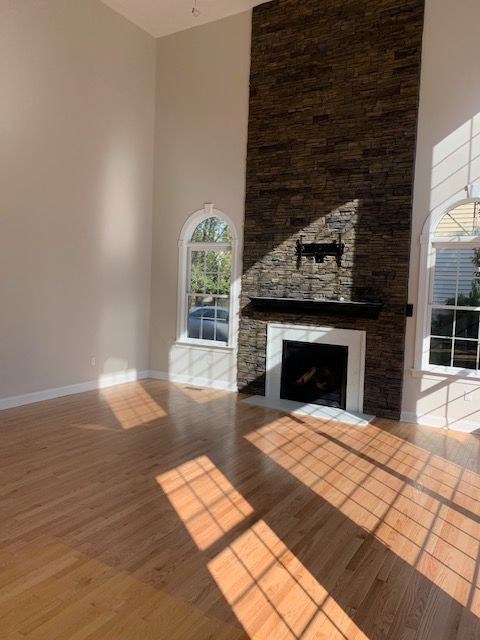 Empty living room with wood floors, fireplace, and two arched windows. Sunlight streams across the floor.