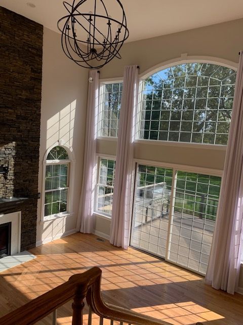 Two-story living room with large windows, light curtains, and a stone fireplace. Sunlight streams onto wooden floors.