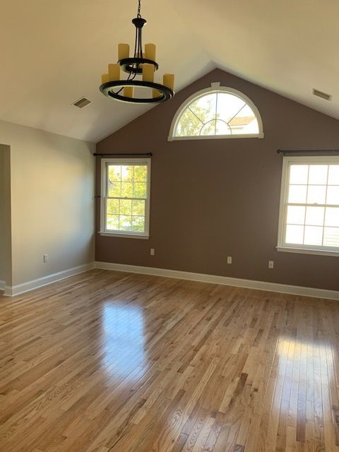 Empty room with wood floors, brown accent wall, arched window, and chandelier.