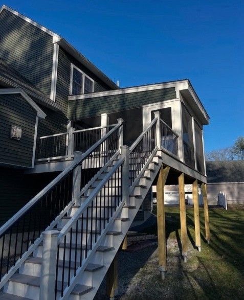 A two-story house with an elevated screened-in porch, accessed by stairs. Green siding, black railings, and a blue sky.