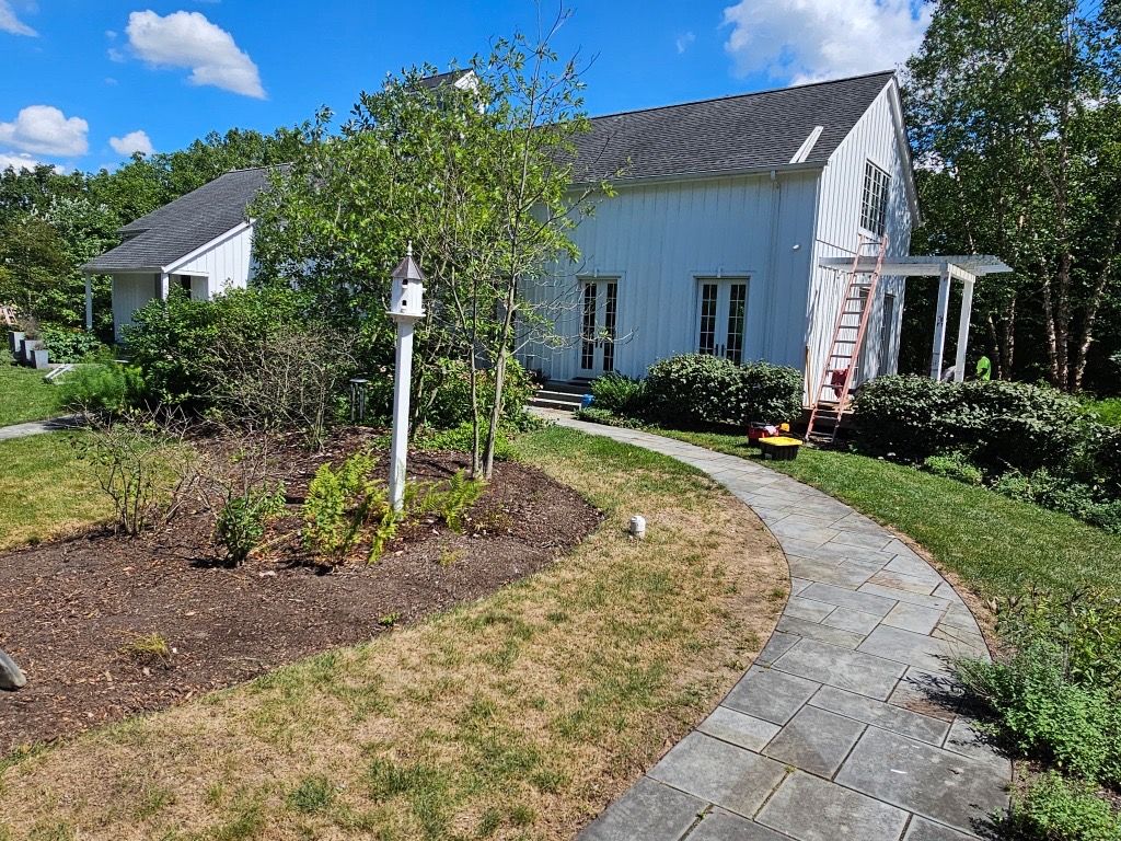 White house with gray roof, a stone path, and a lush garden under a blue sky.
