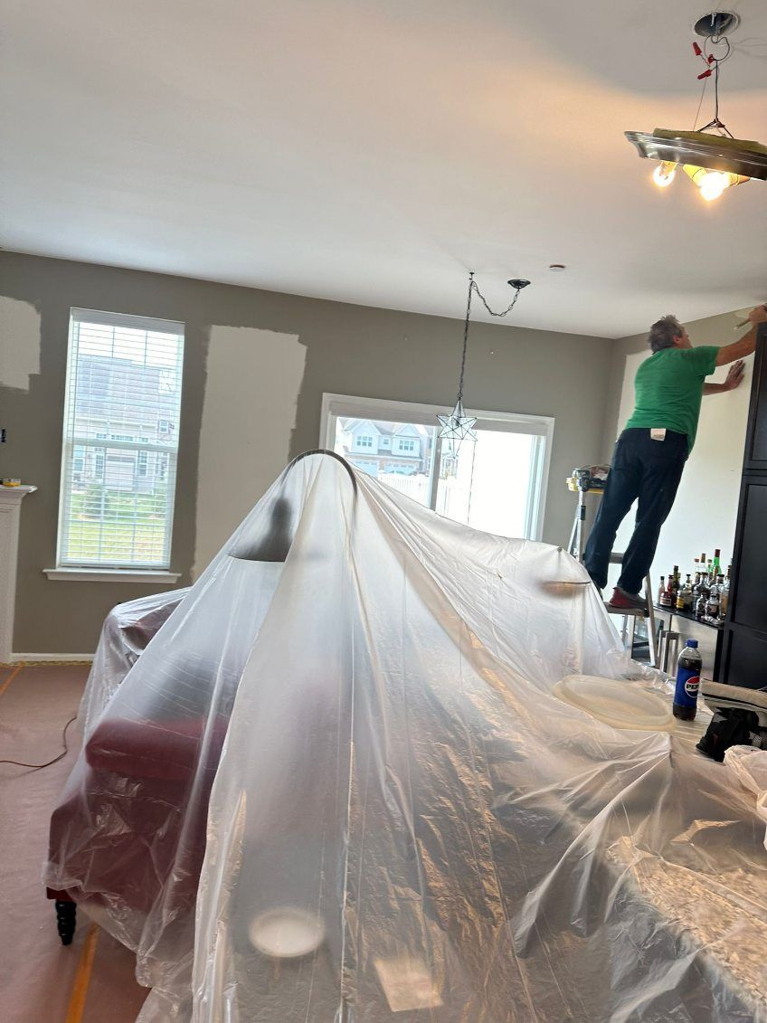 Person painting ceiling, standing on ladder in living room, furniture covered in plastic.