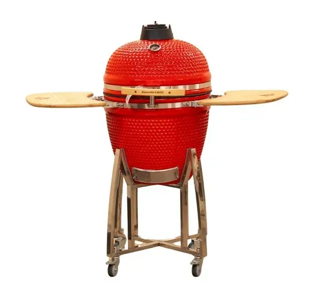 Red ceramic kamado grill on a metal stand with wooden side shelves, isolated on white