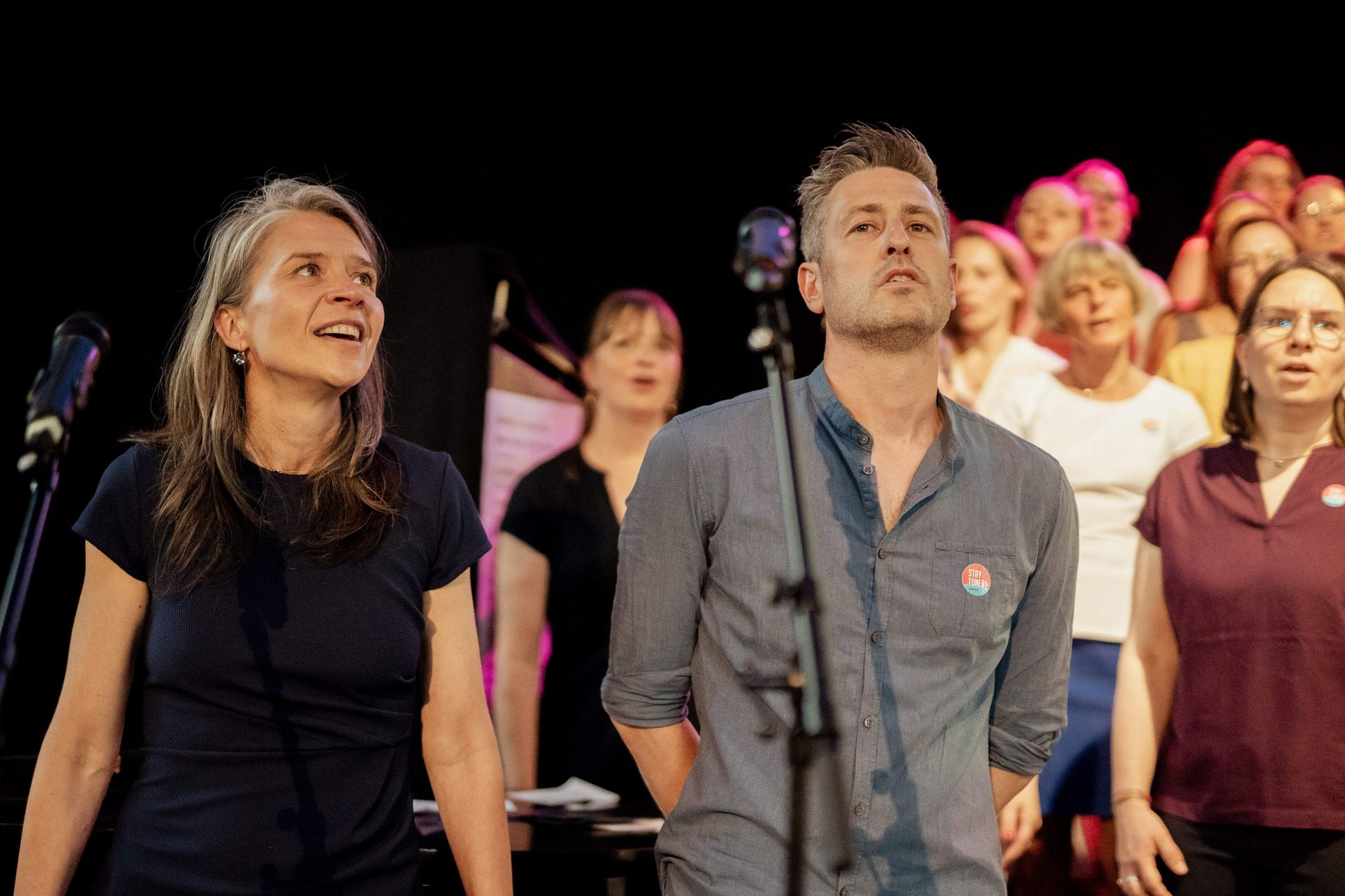 Eine weiblich und männlich gelesene Personen singen zusammen auf einer Bühne, im Hintergrund ein Chor.