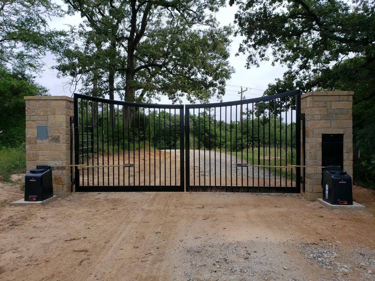 Black metal gate between brick pillars at a dirt driveway entrance, with trees behind.