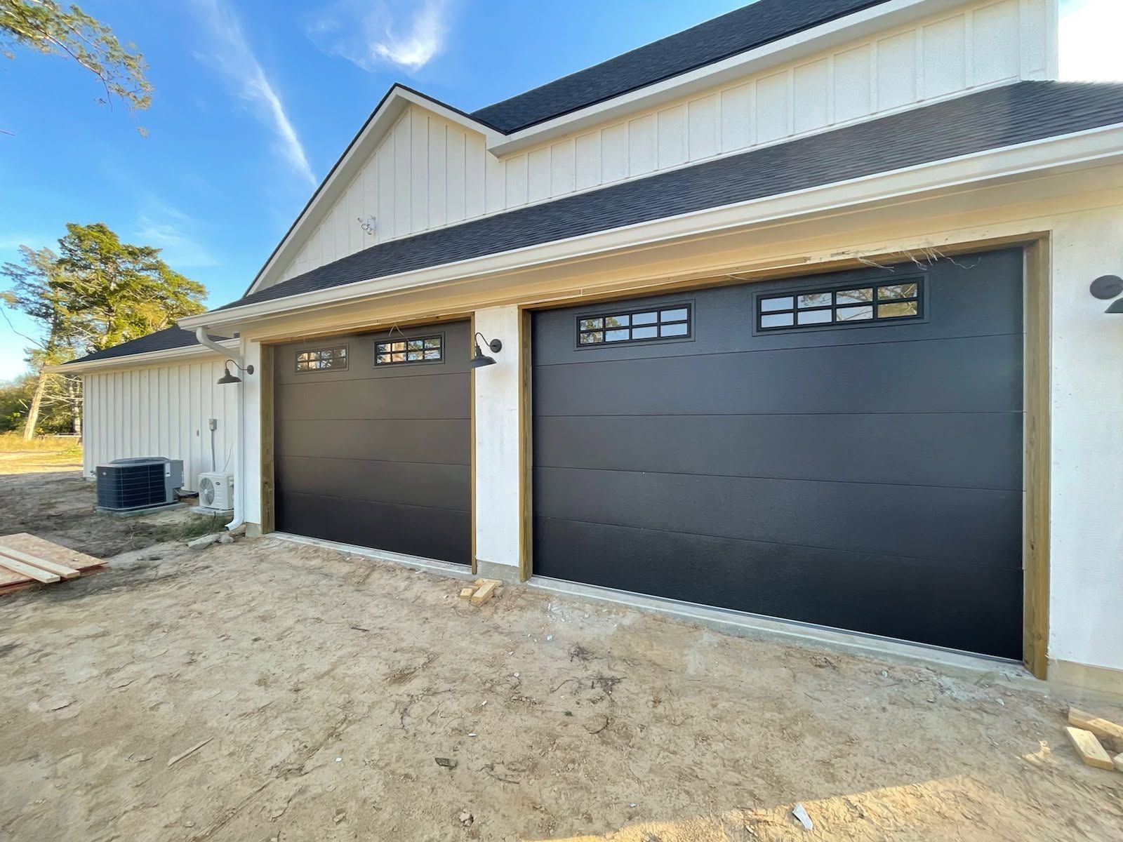 New home exterior with two dark garage doors and unfinished dirt driveway under a clear blue sky