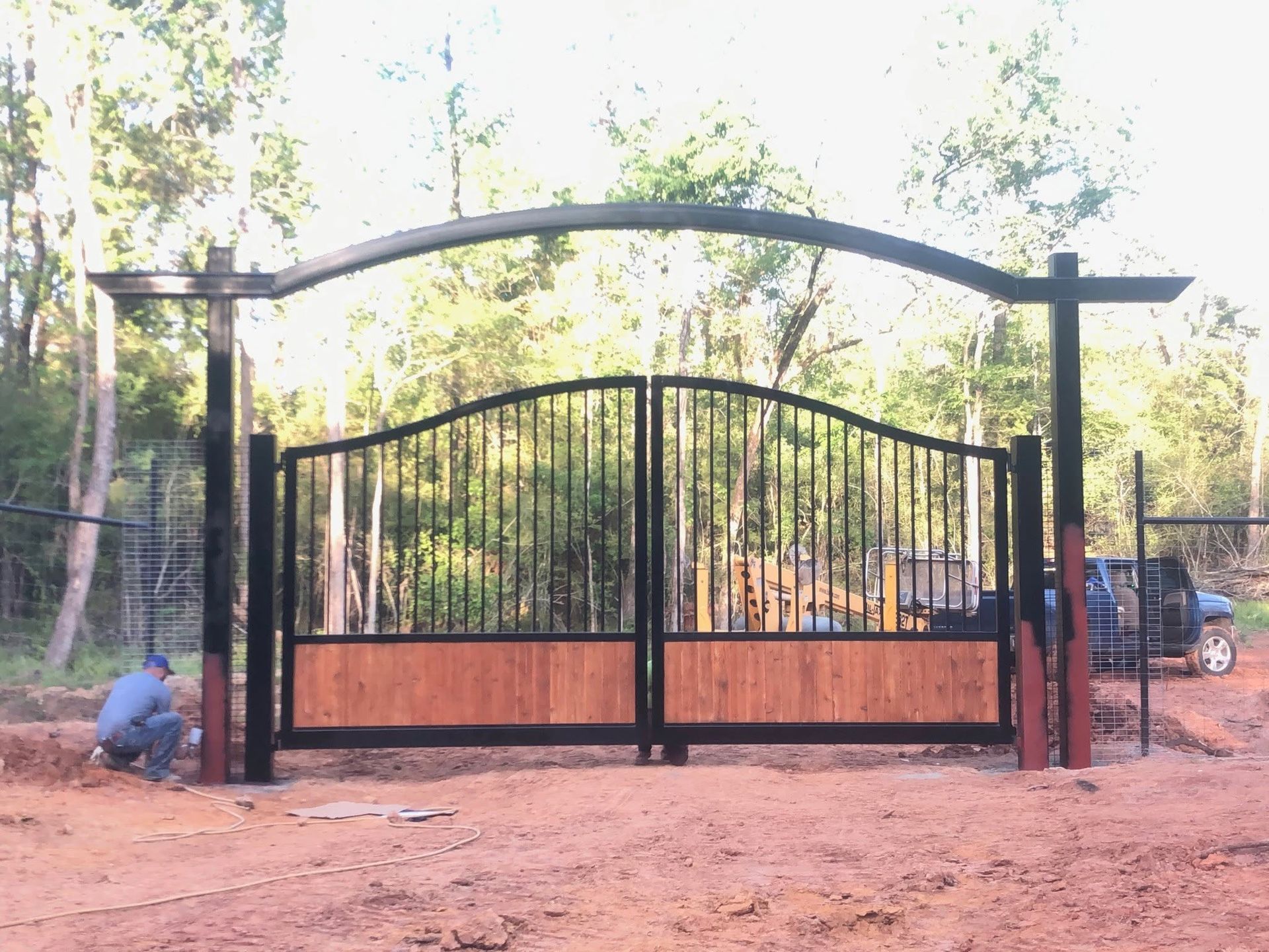 Black metal gate and fence at a wooded driveway entrance, with a person crouching on the left.