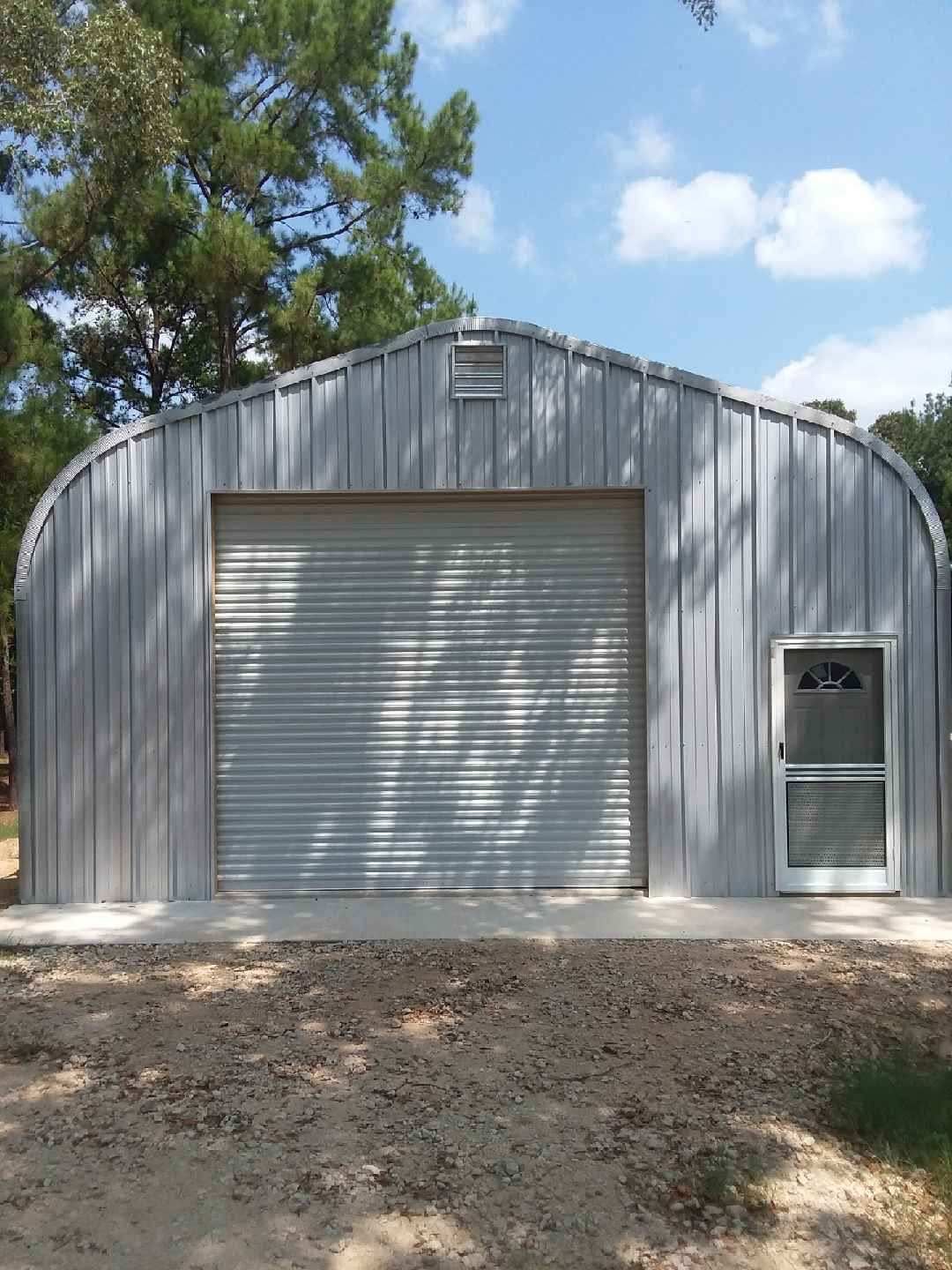 Metal garage with roll-up door and side entry door under a blue sky, surrounded by trees and dirt ground