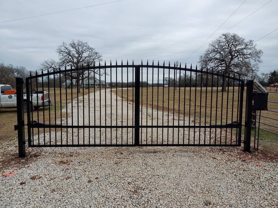 Black wrought-iron driveway gate at a gravel entrance on a cloudy day