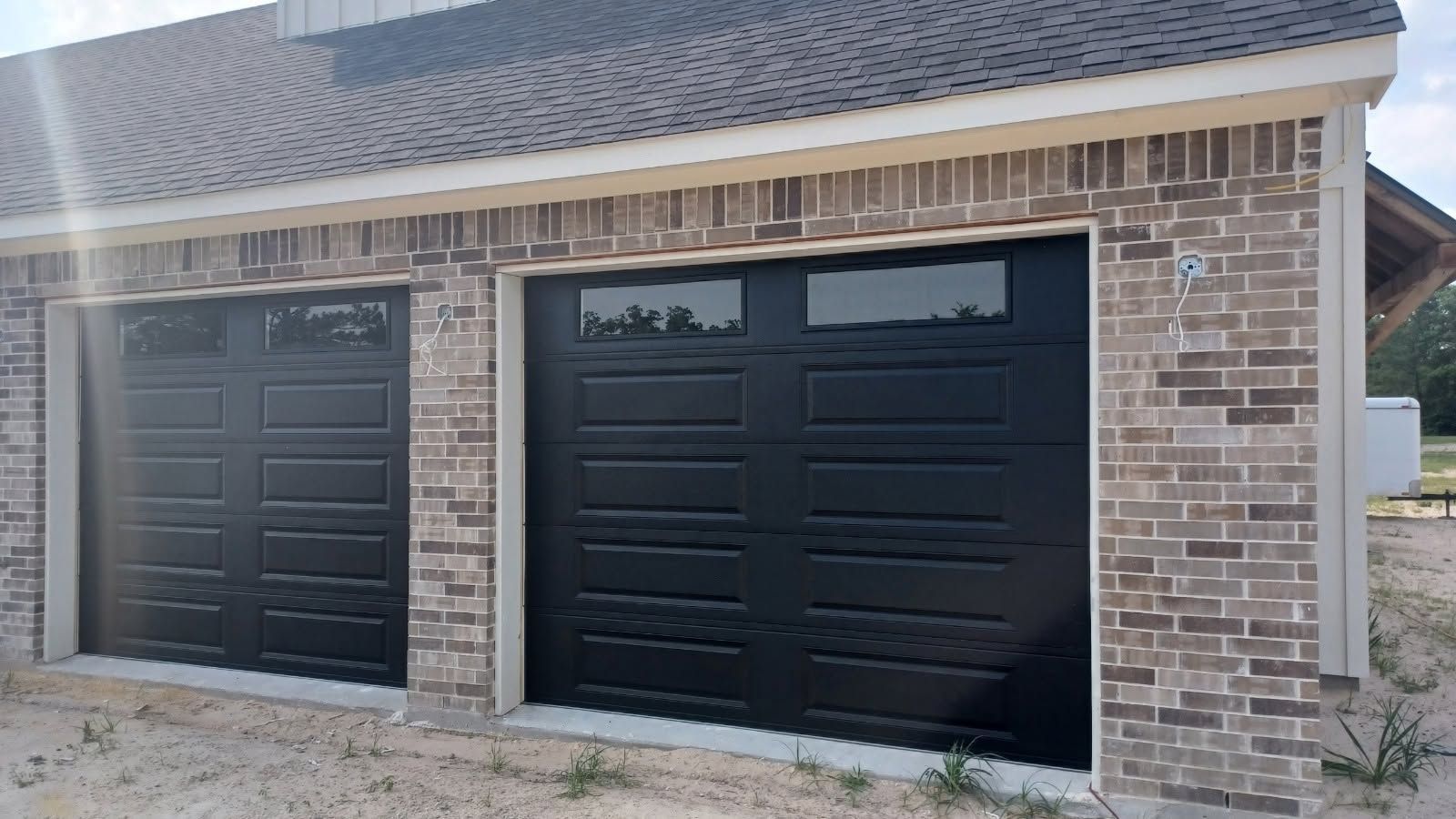 Brick garage with two black paneled doors and a shingled roof.