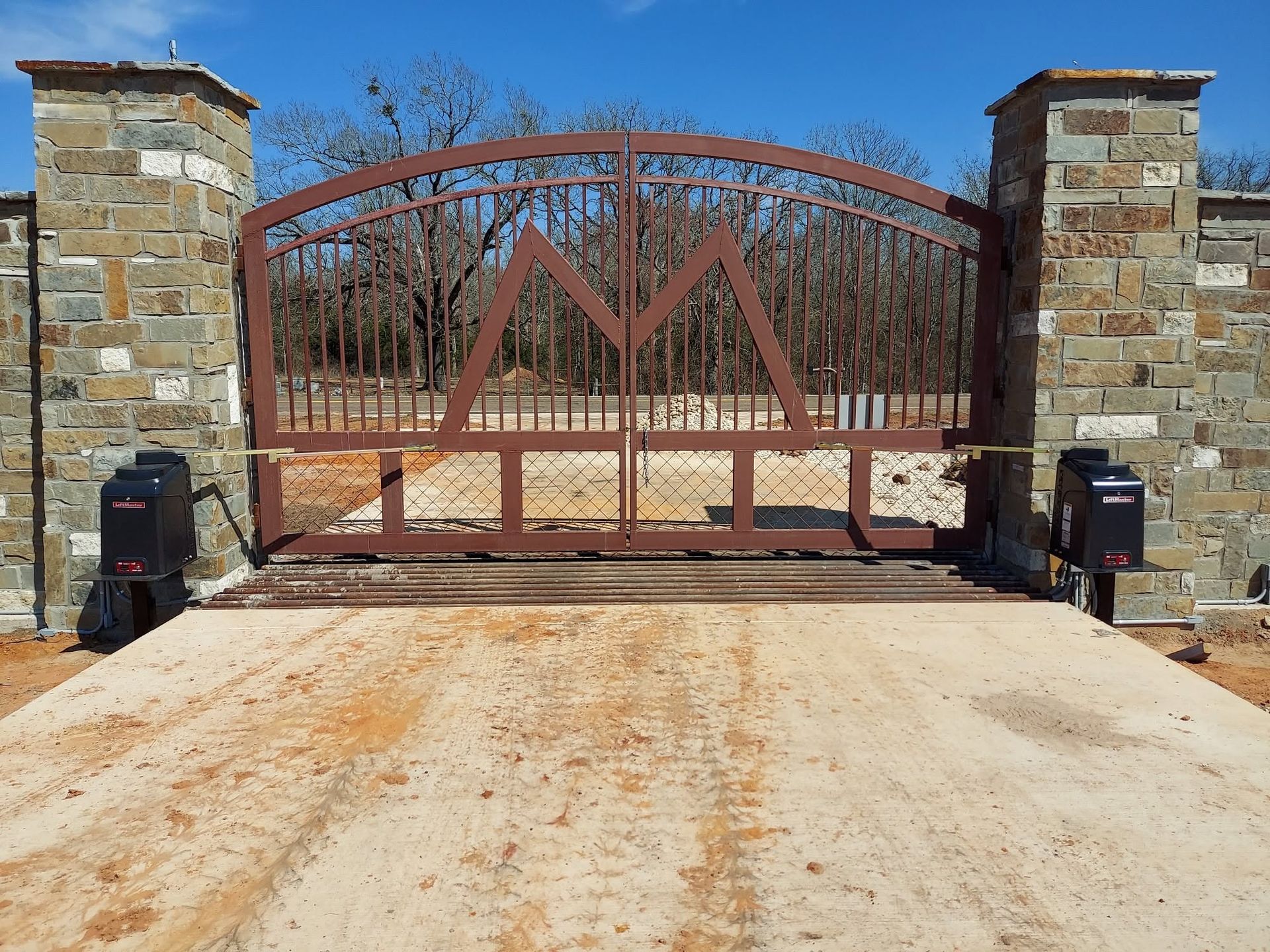 Closed rust-colored metal gate between stone pillars on a driveway under a clear blue sky