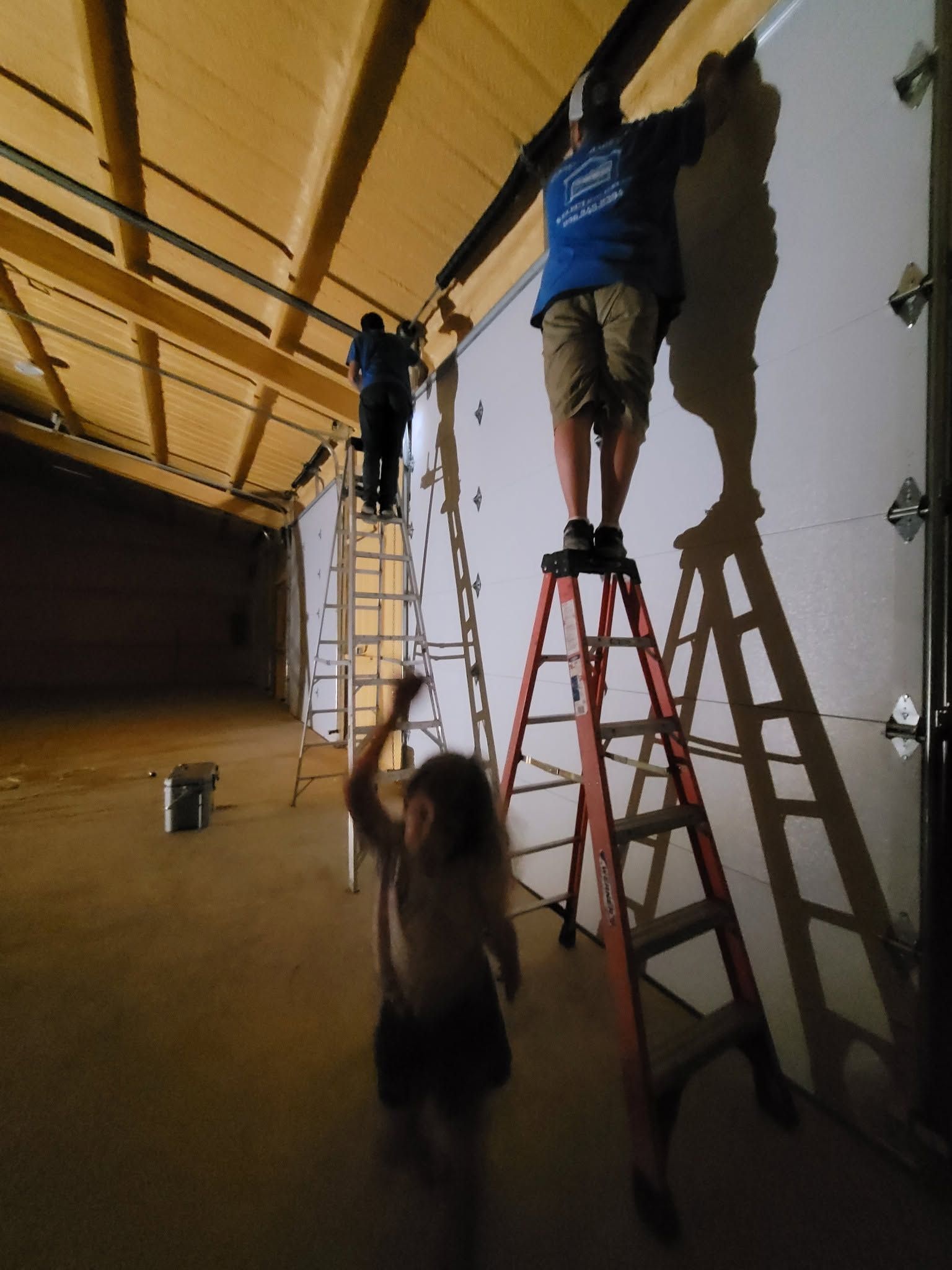 People on ladders painting a wall in a large indoor space with a child nearby