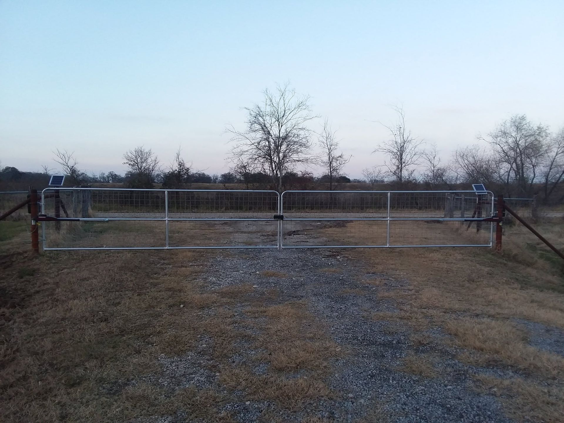 Closed metal gate across a gravel driveway in a rural field at dusk