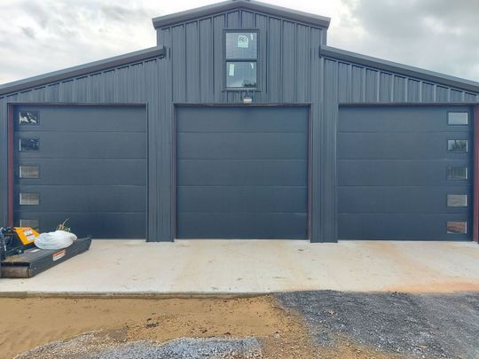 Dark gray metal garage with three black doors, a small upper window, and a concrete driveway.