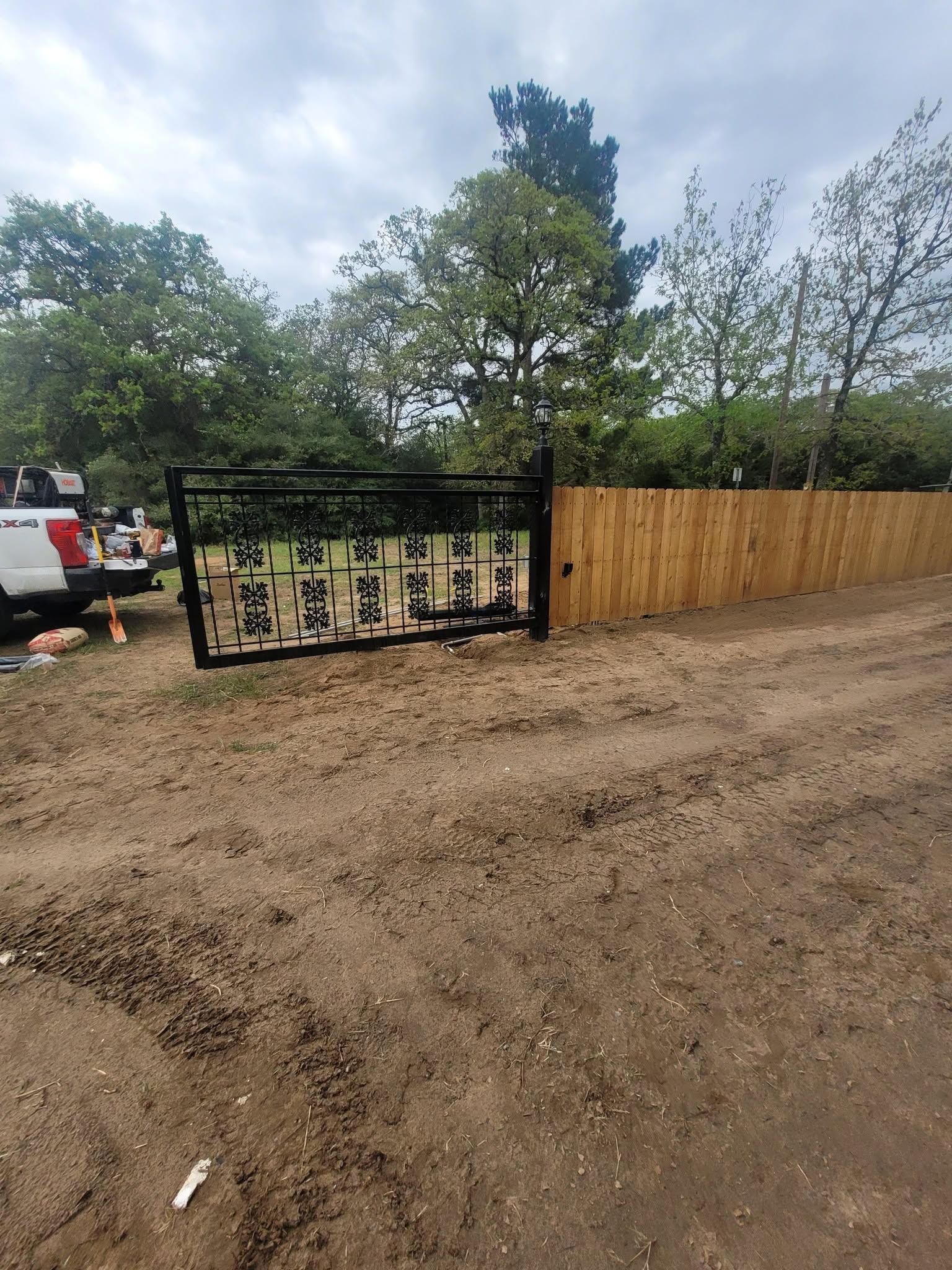 Dirt lot beside a wooden fence, with a black metal sign frame and trees under a cloudy sky.
