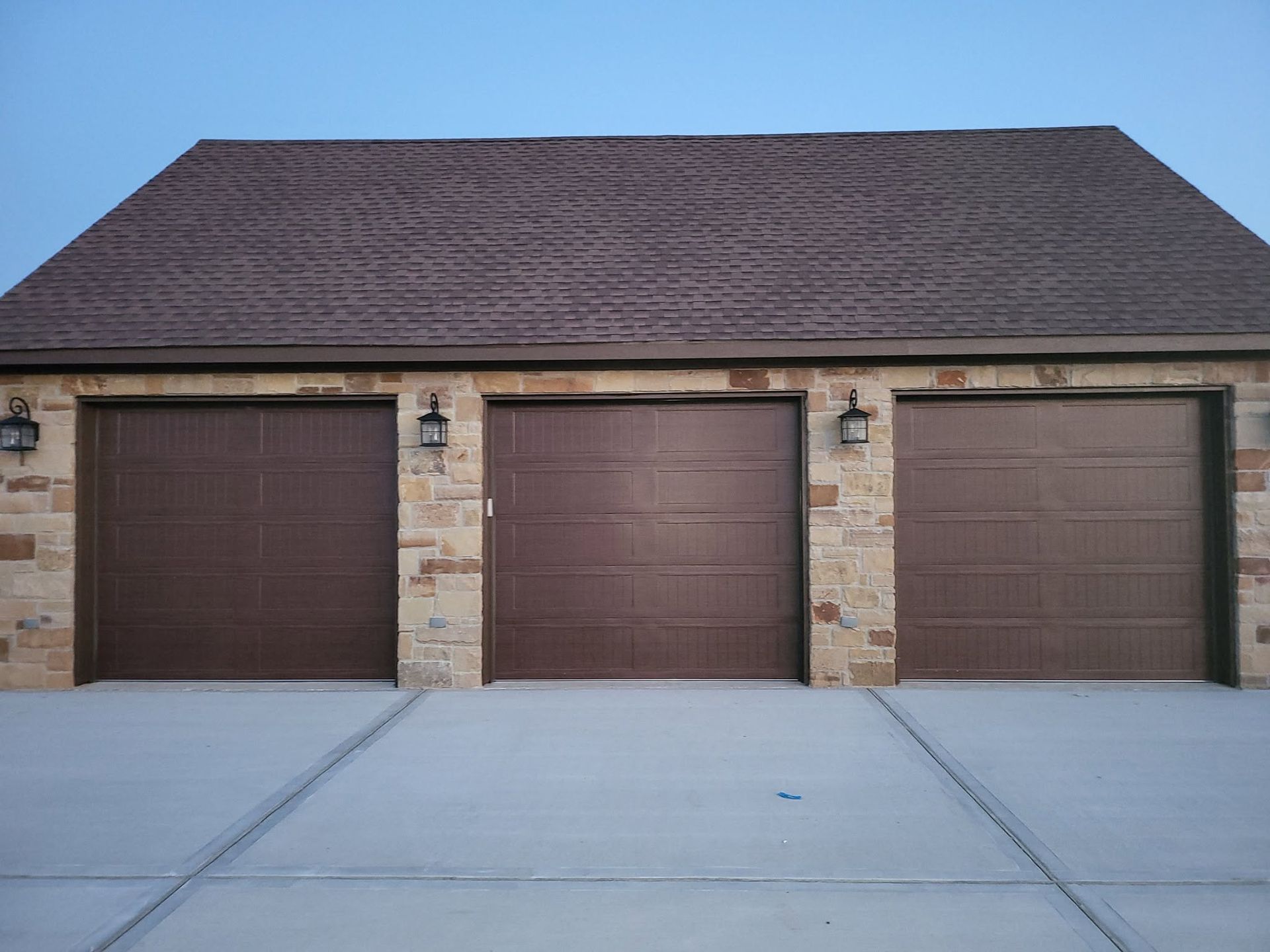 Three brown garage doors on a stone house with a gray driveway under a clear blue sky
