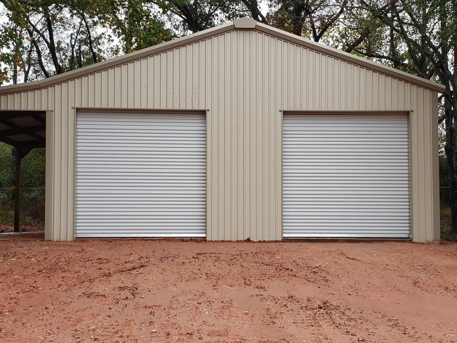Two white roll-up garage doors on a beige metal shed with a red dirt foreground.