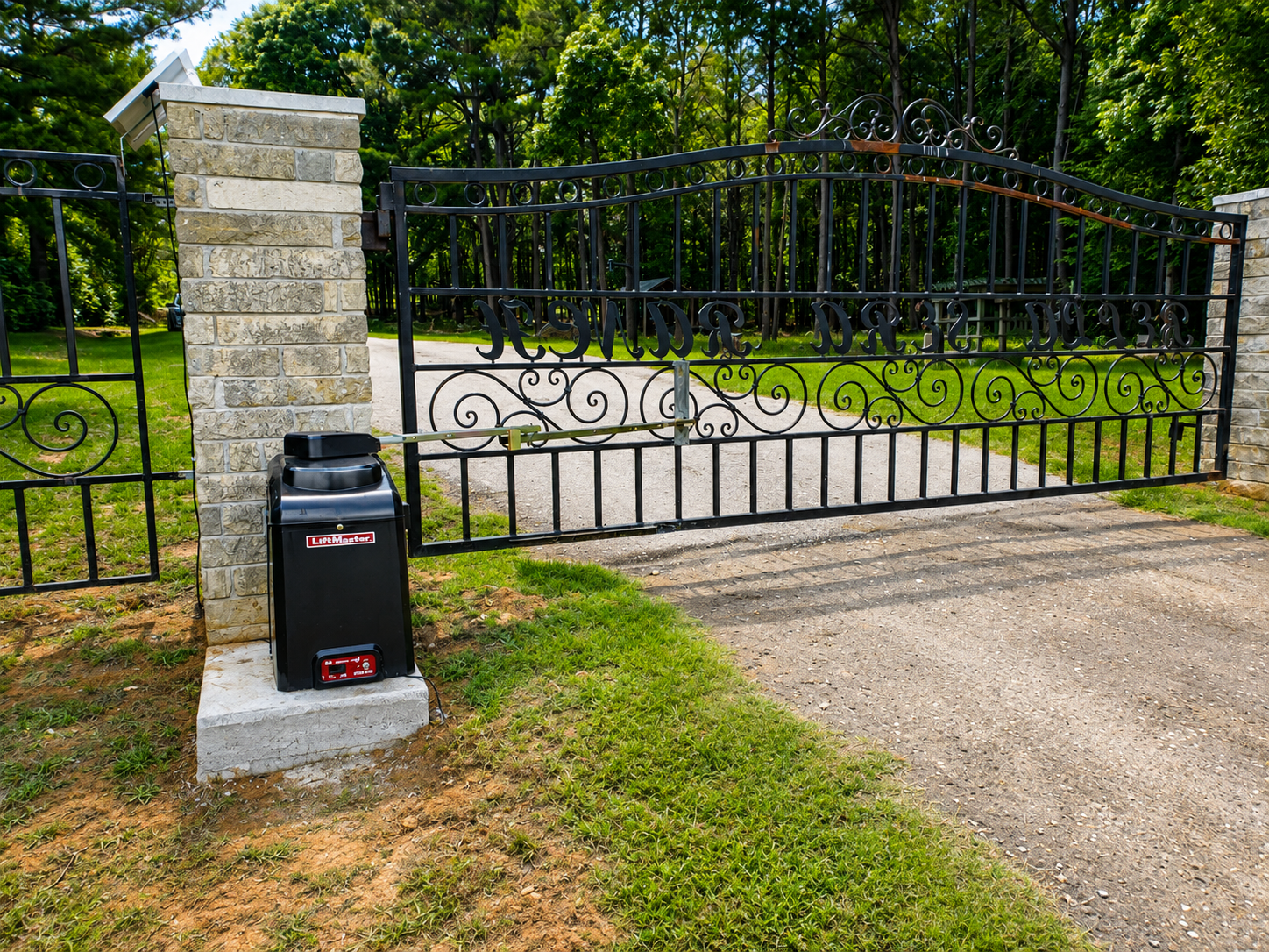 Black iron gate beside a stone pillar, with a black mailbox on a grassy driveway.