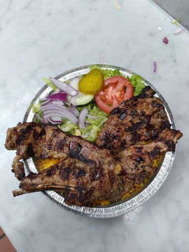 Grilled meat with rice and salad in a silver container. Includes tomato, onions, lettuce, and a pickle.
