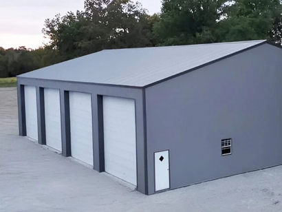 Gray metal garage with four bays and a small white door, set against a green tree line and overcast sky.