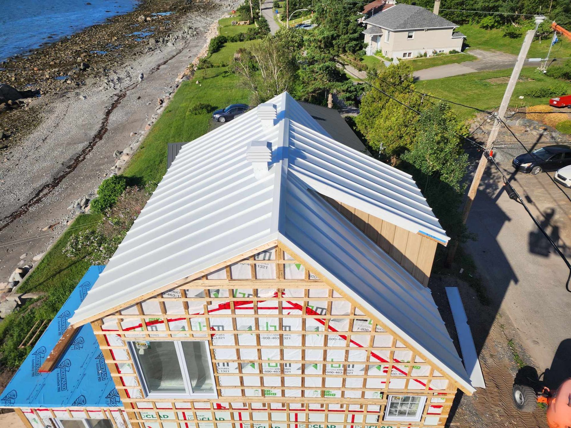 Vue aérienne d'une maison dont l'extérieur est partiellement construit ; toit en métal blanc, ossature en bois, bâche bleue.
