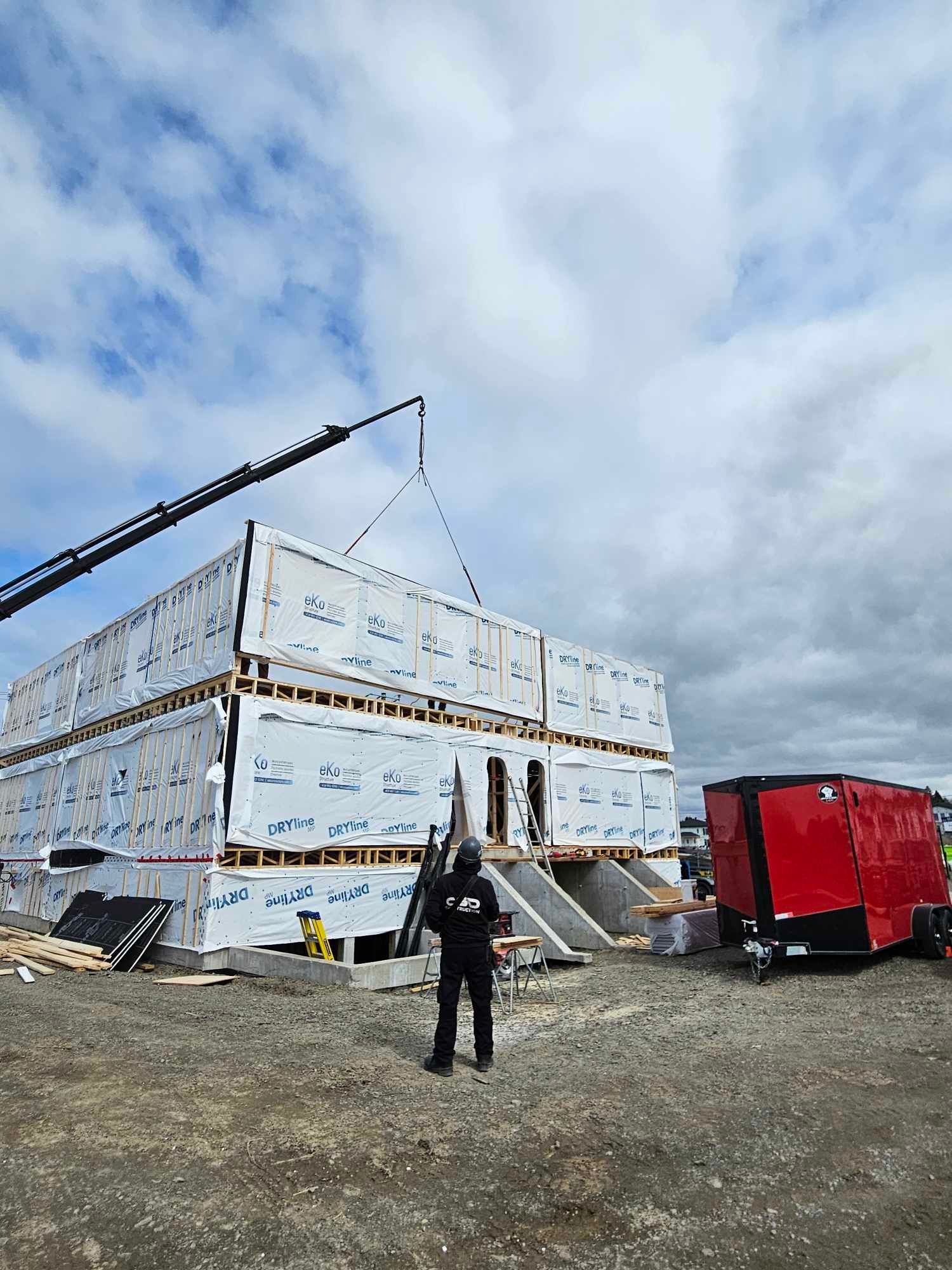 Chantier : une grue soulève un module de construction, un ouvrier se tient à proximité. Remorque rouge à droite, ciel bleu.