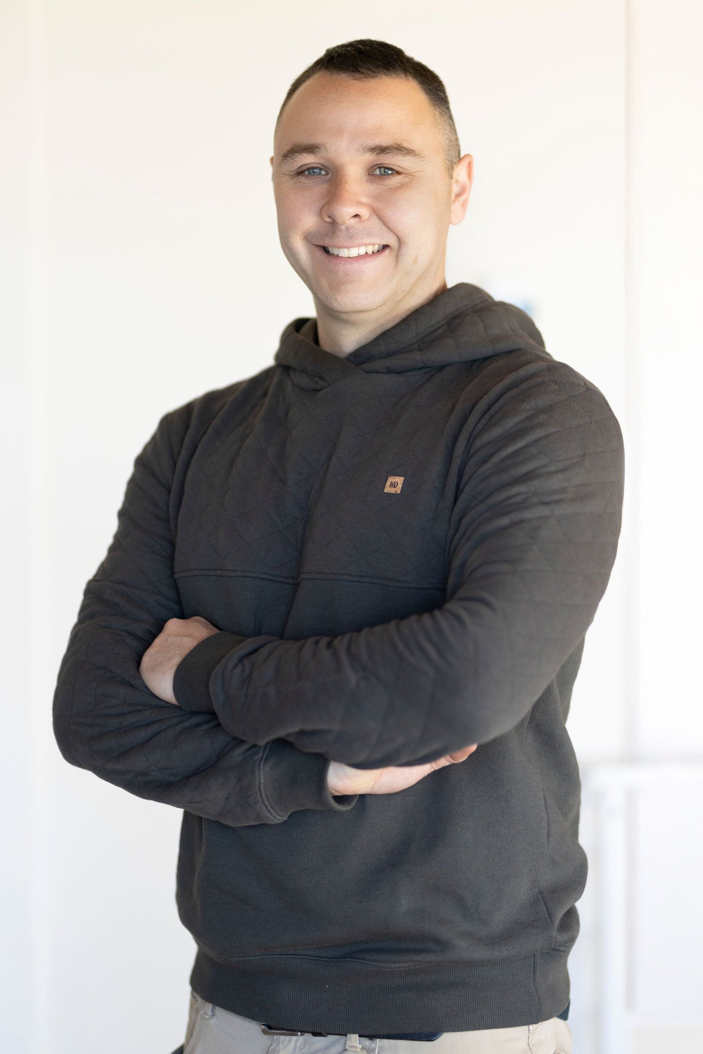Un homme en sweat-shirt gris, les bras croisés, souriant, se tient devant un mur blanc.