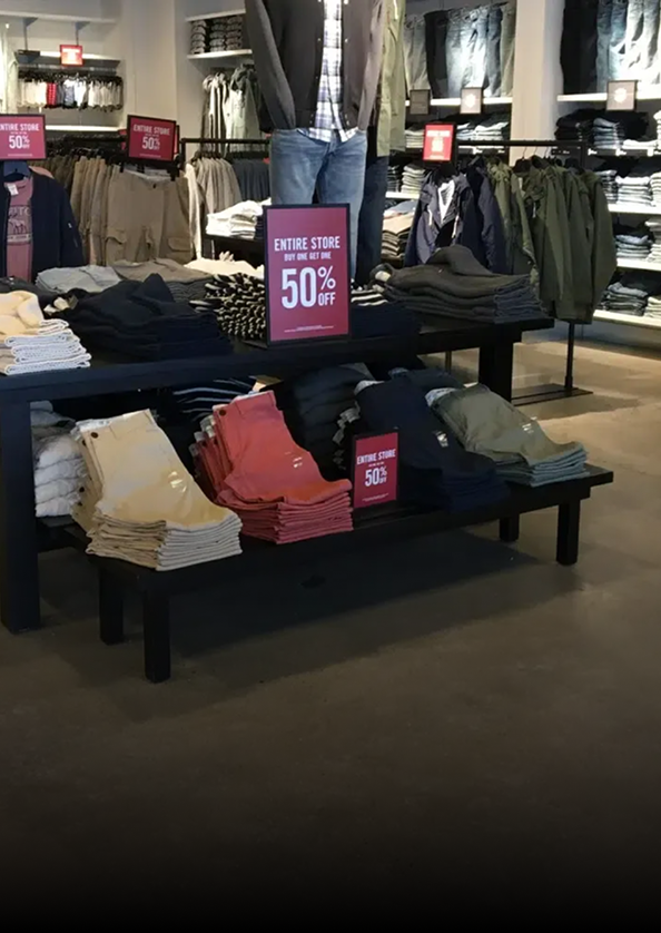 Clothing store display with folded jeans and shirts on tables, racks behind, and a 50% off sale sign.