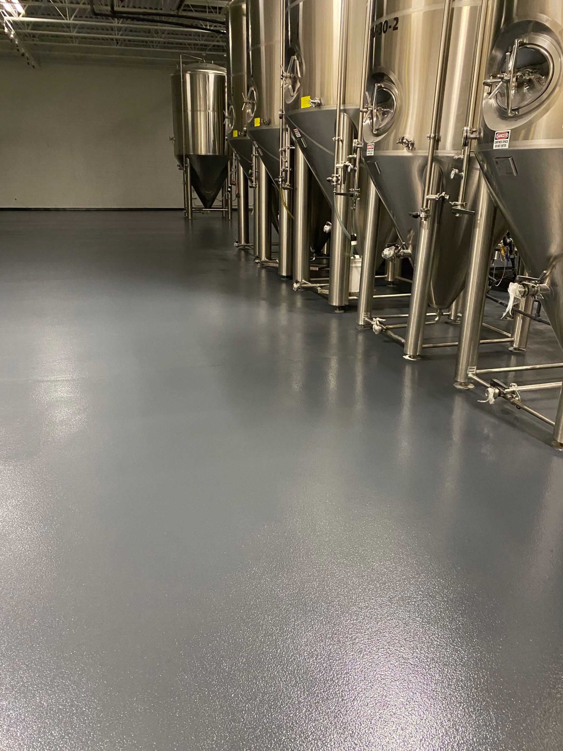 Industrial brewery floor with stainless steel tanks and pipes along a gray epoxy surface