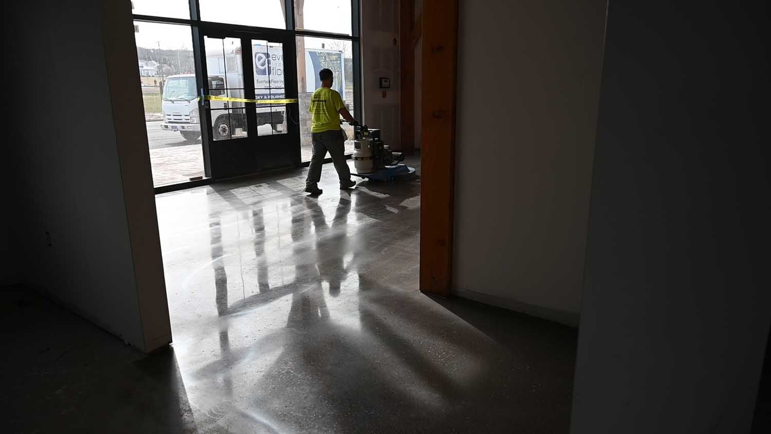 Worker in a yellow jacket mopping a shiny floor inside a building near glass doors.